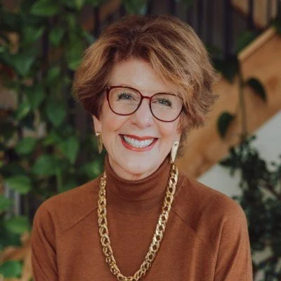 A smiling woman with short, wavy hair, glasses, wearing a brown turtleneck, gold chain necklace, and earrings, with a background of plants and wooden structures.