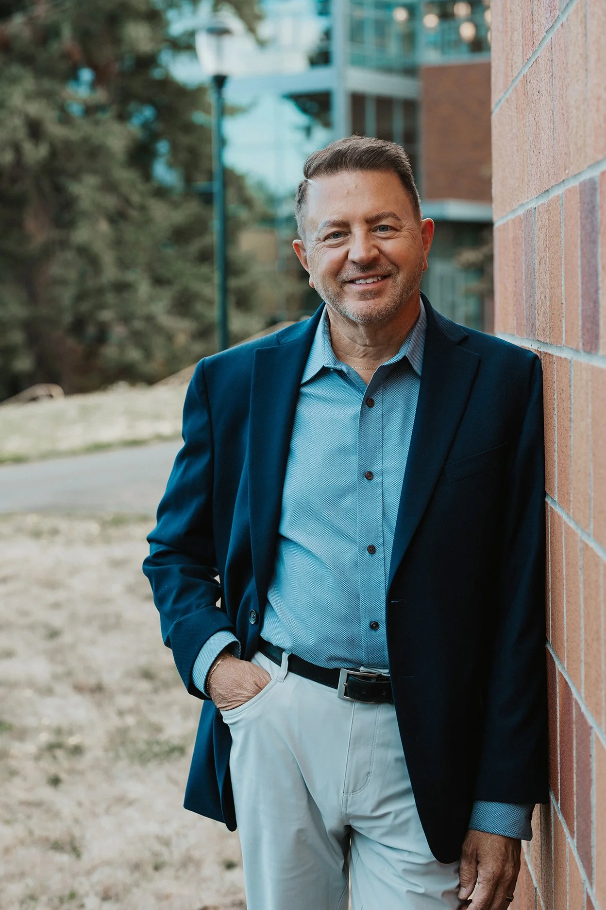 A man in a navy blazer and light blue shirt smiling while leaning against a brick wall outdoors.