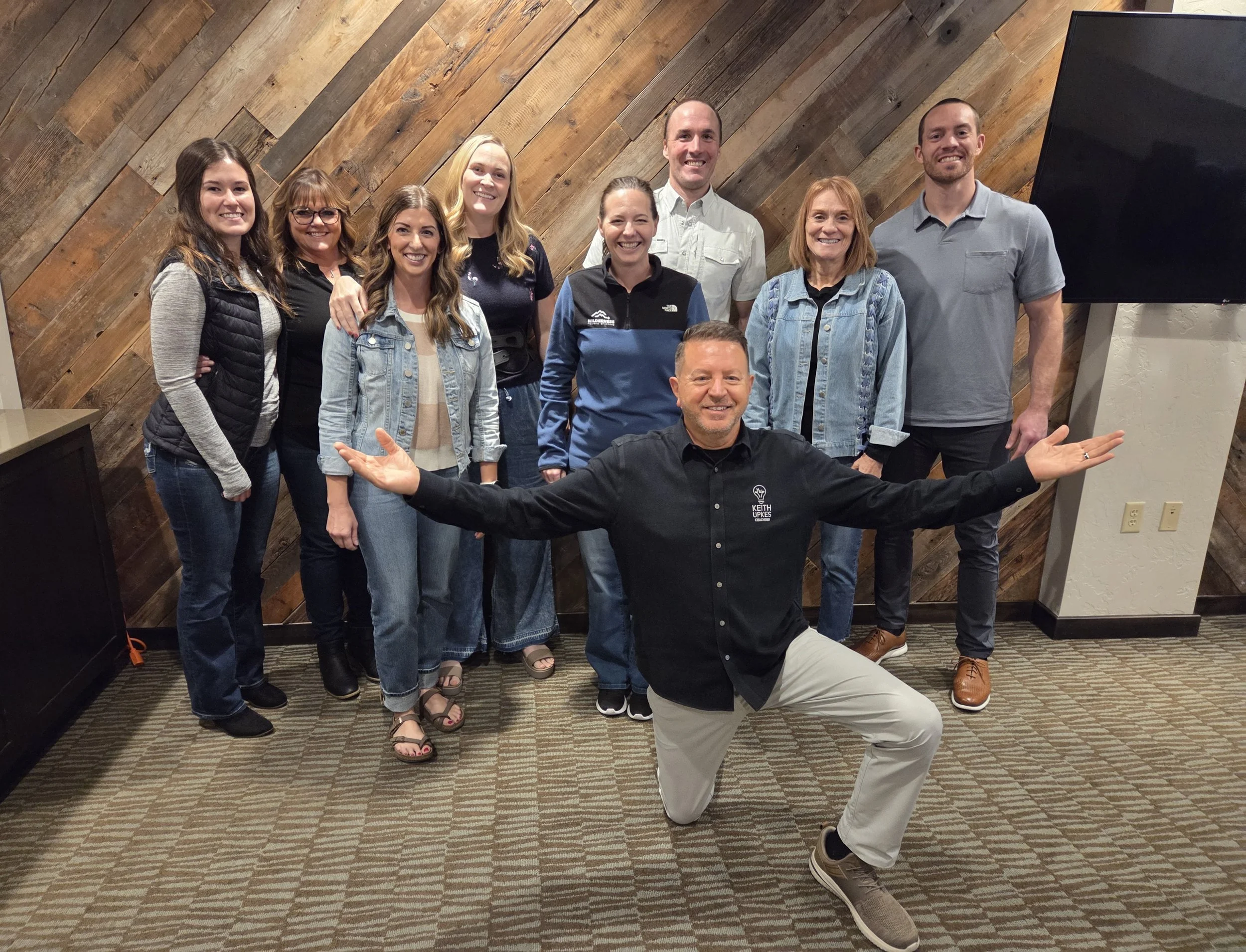 A group of ten people posing indoors in front of a wooden wall, with a man kneeling in front with arms outstretched, all smiling.