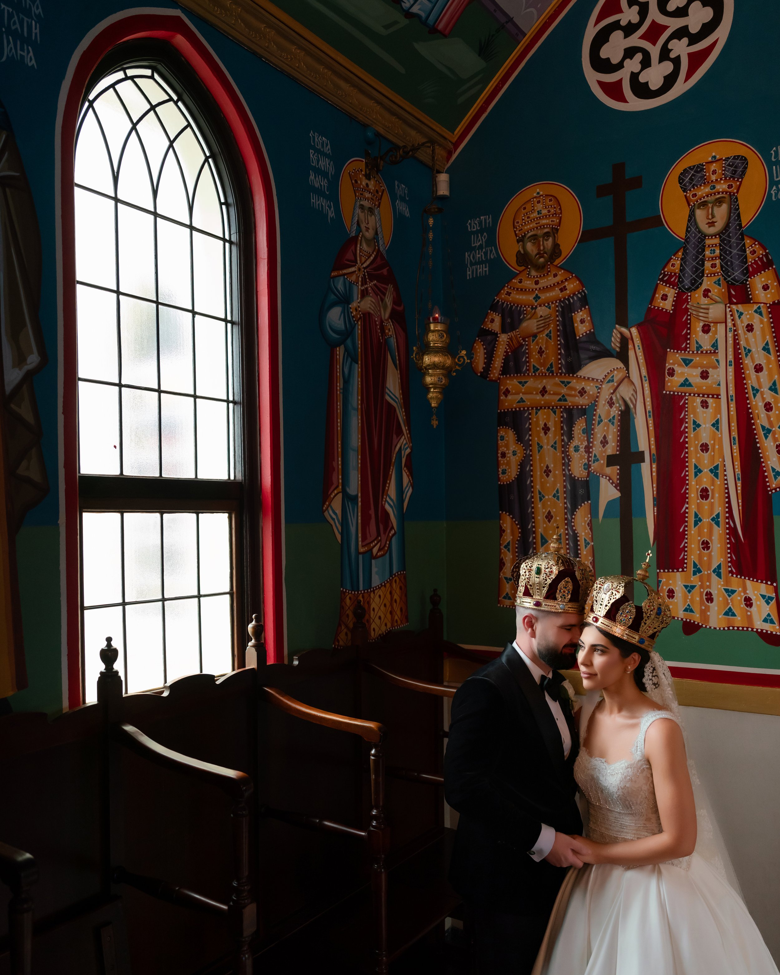 A bride and groom in wedding attire standing close together inside a church, with religious iconography, stained glass windows, and wooden pews visible in the background.