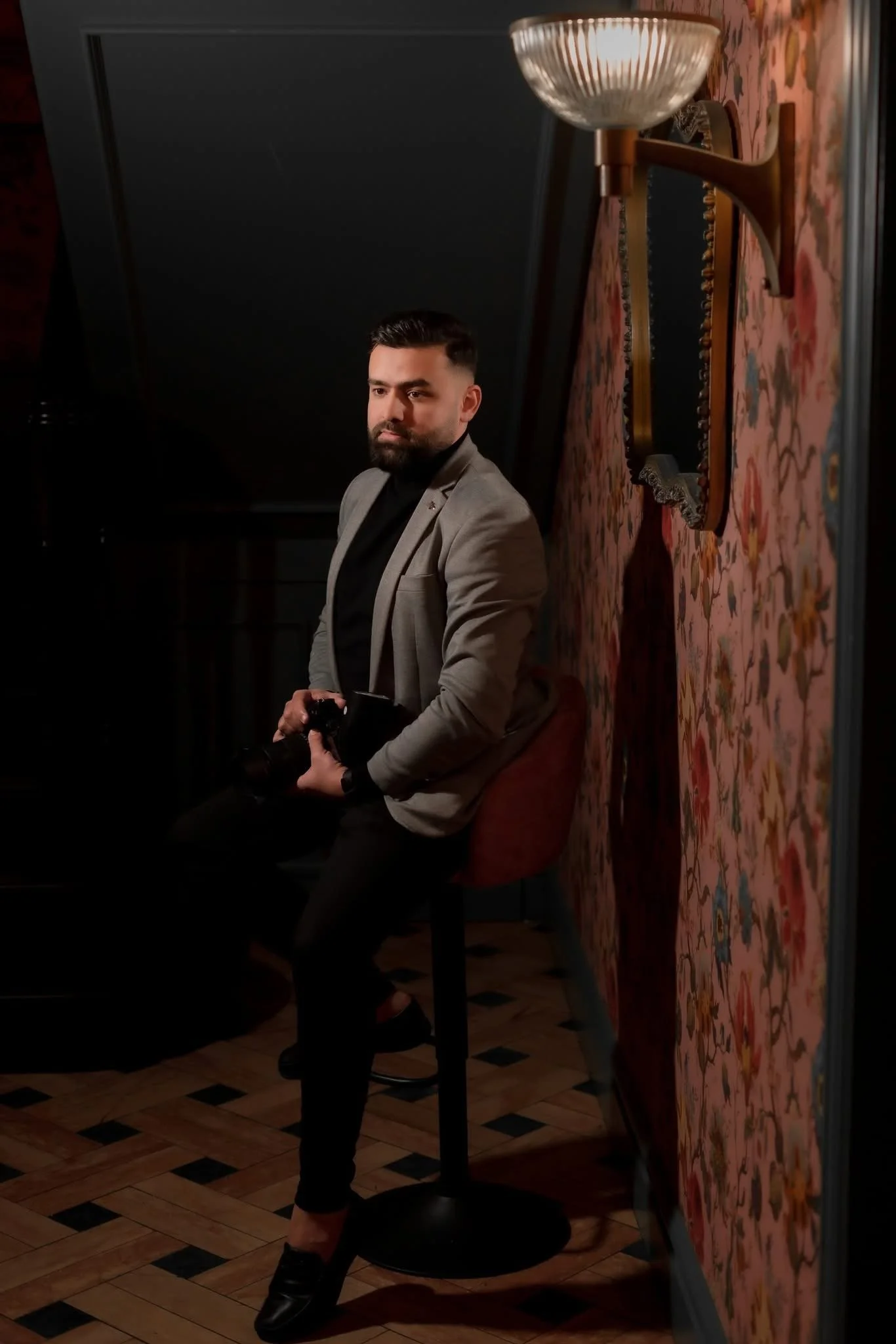 A man with a beard and styled hair, dressed in a gray blazer and black shirt, sitting on a bar stool against a floral wallpapered wall, holding a camera in his lap, in a dimly lit room with a patterned wooden floor and a wall-mounted lamp above.