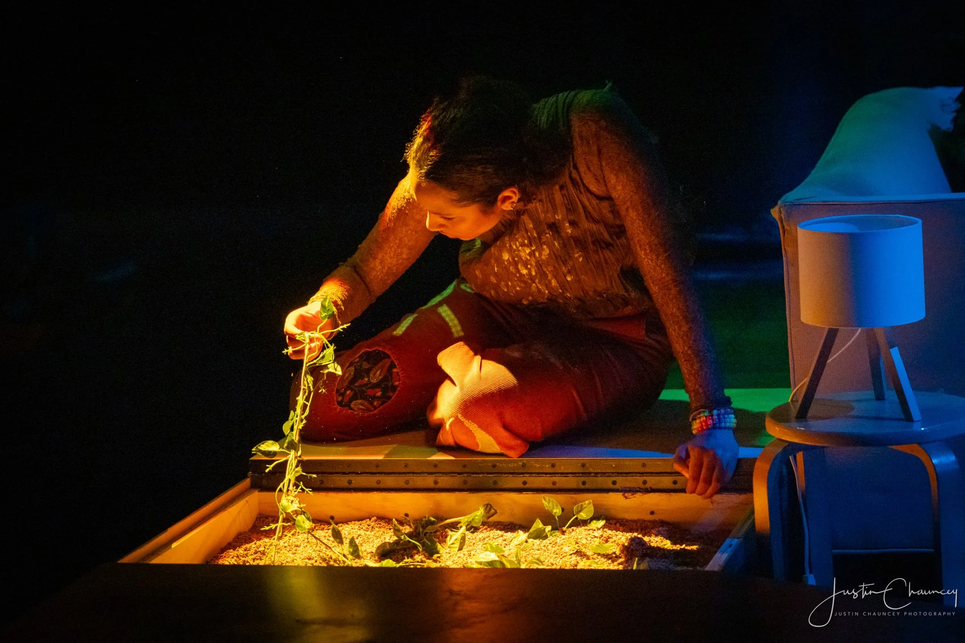 A person with long dark hair, wearing a colorful long sleeve top and patterned patchwork pants, is kneeling on a small platform in a dark room. They are tending to small green plants growing in soil illuminated by warm lighting. There is a blue and orange lamp on a small table nearby.
