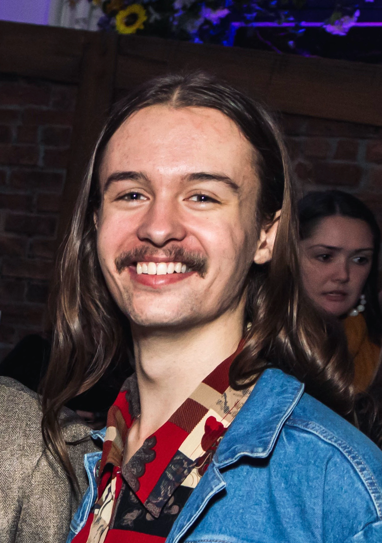 A young man with long brown hair, mustache, and goatee, smiling, wearing a denim jacket and patterned shirt, at a social gathering.