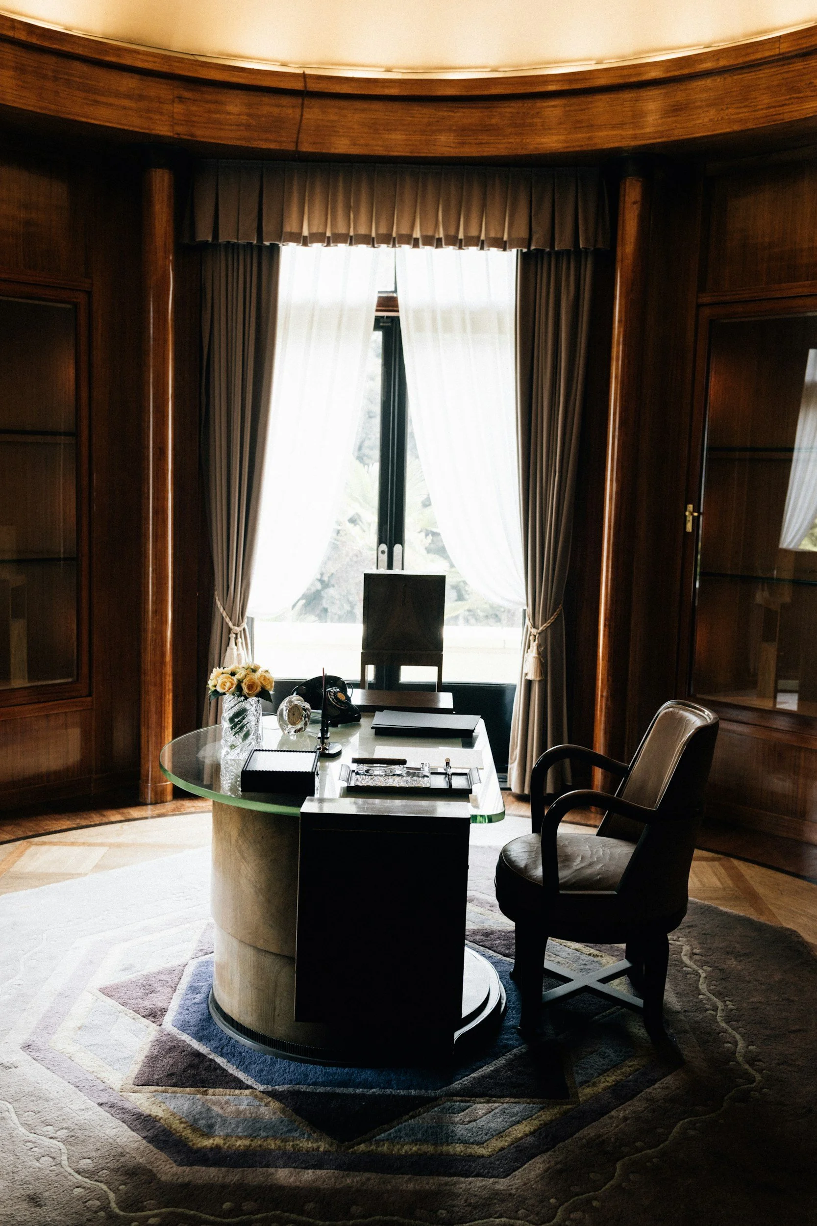 A wood-paneled office with a large window covered by sheer white and heavier brown curtains, a glass-topped round desk with items like a vase with flowers, electronic devices, and a black box, and two upholstered chairs surrounding the desk. The room has a decorative carpet and natural light.