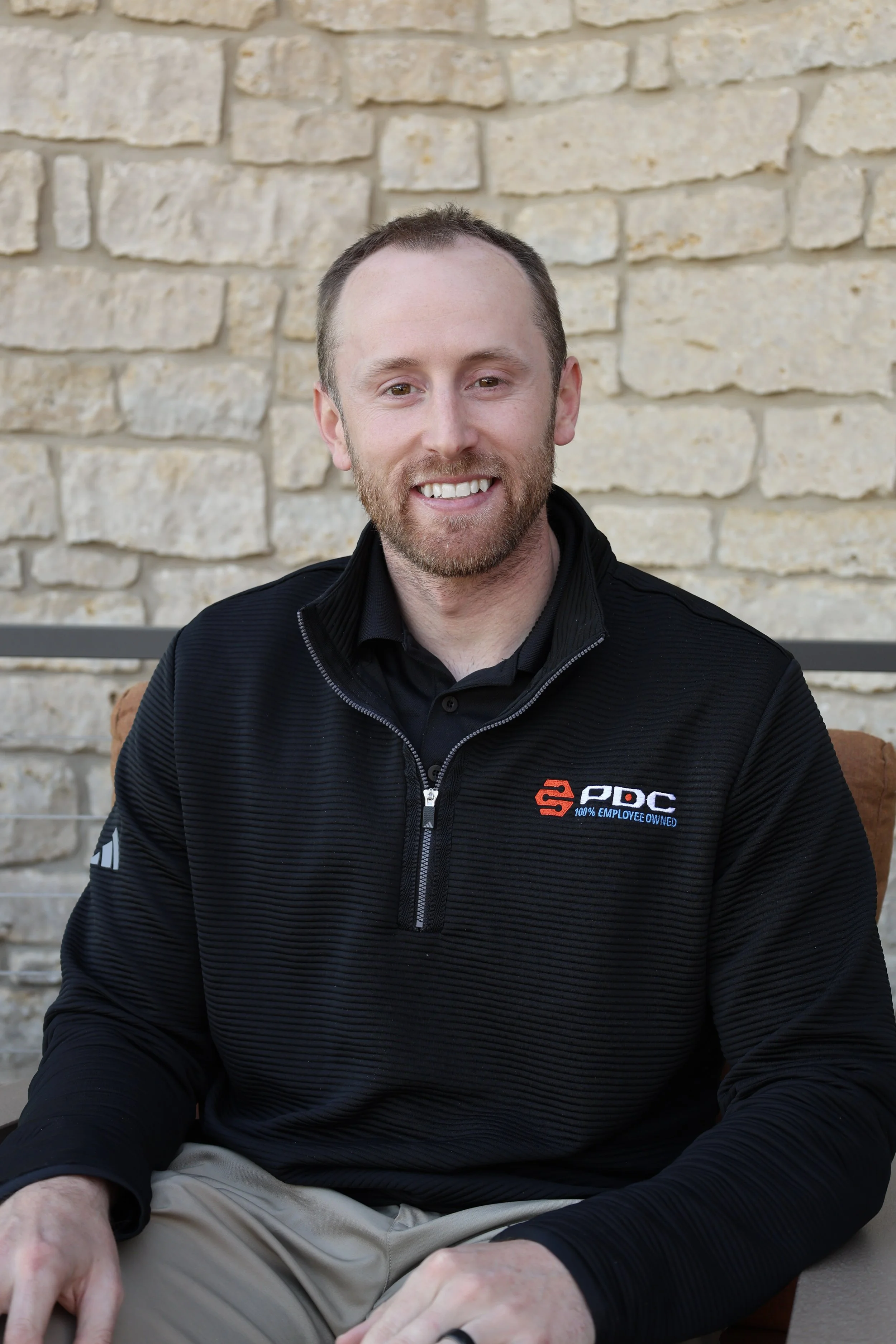 Headshot of a smiling man with a beard, wearing a black jacket with a logo, sitting in front of a stone wall.