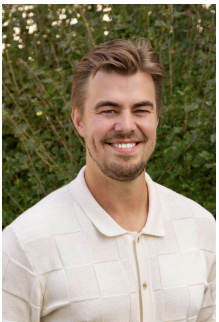 Young man with light brown hair and beard smiling outdoors, wearing a white collared shirt, with green foliage in the background.