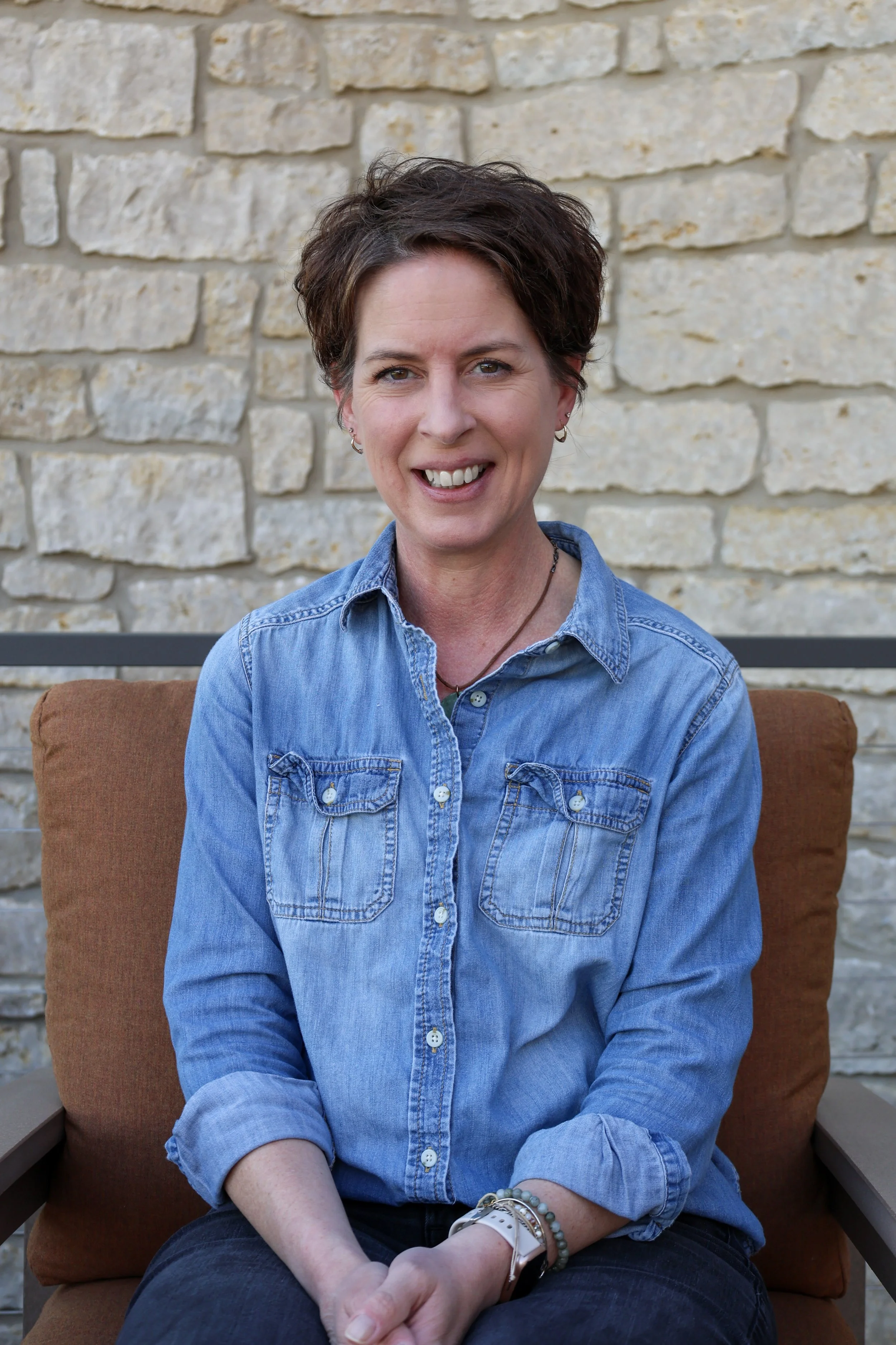 A woman sitting on a brown cushioned patio chair, smiling, in front of a beige brick wall. She has short, dark hair and is wearing a blue denim button-up shirt, dark pants, multiple bracelets, and earrings.