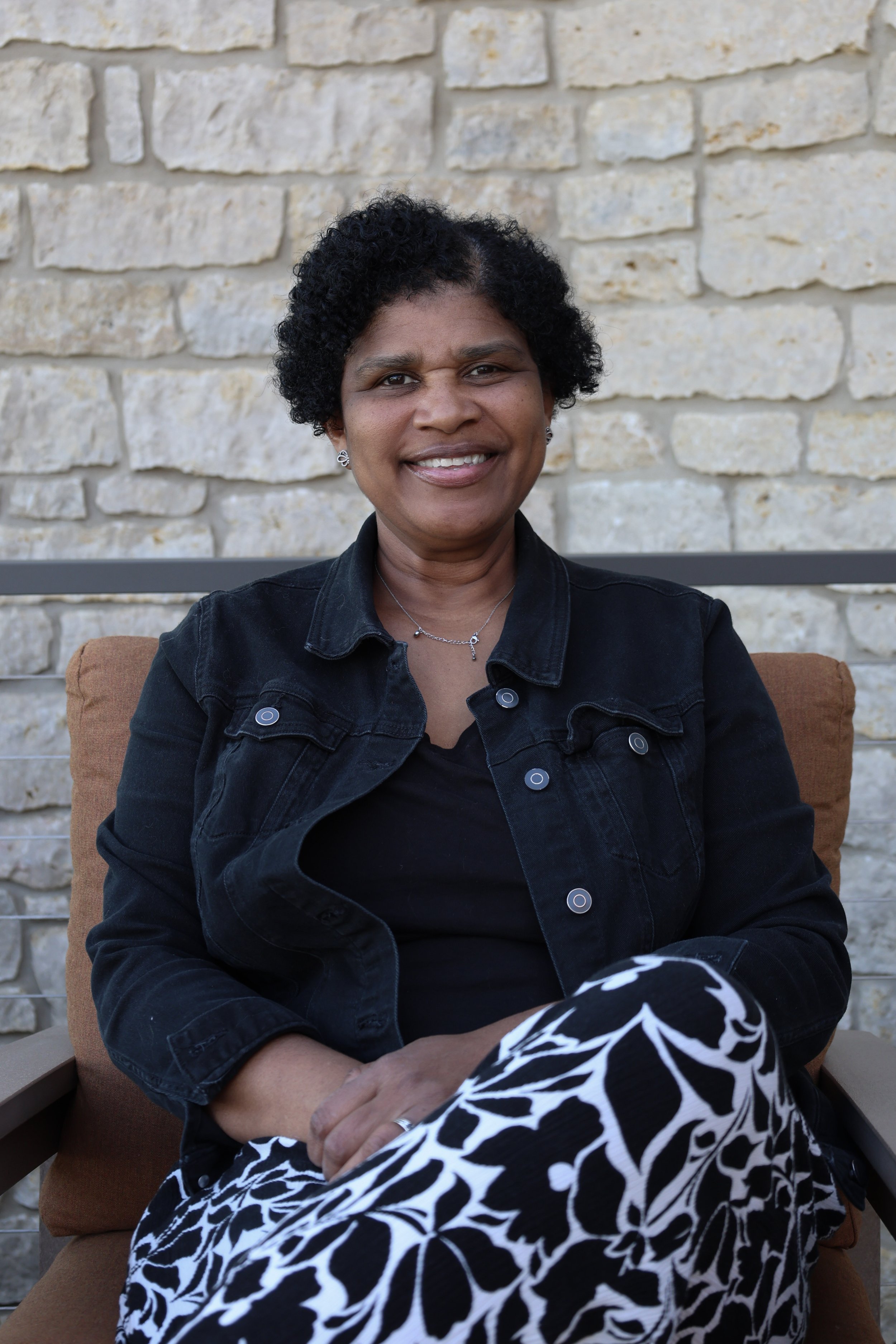 A middle-aged woman with short curly black hair, wearing earrings, a necklace, a black shirt, and a black denim jacket, sitting outdoors on a cushioned seat with a beige and white stone wall in the background.