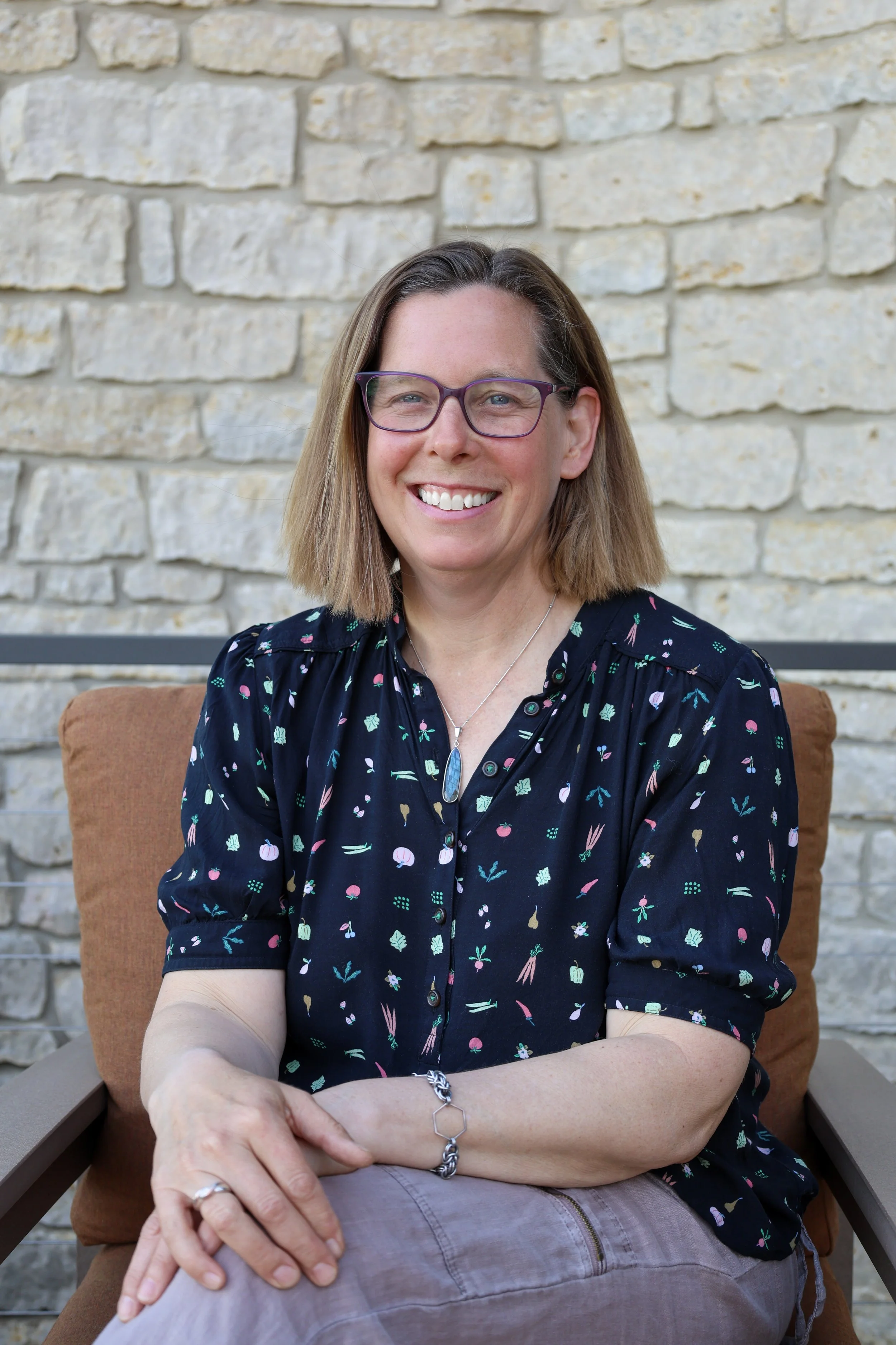Smiling woman with brown hair and glasses sitting outside against a stone wall, wearing a dark floral blouse and silver jewelry.