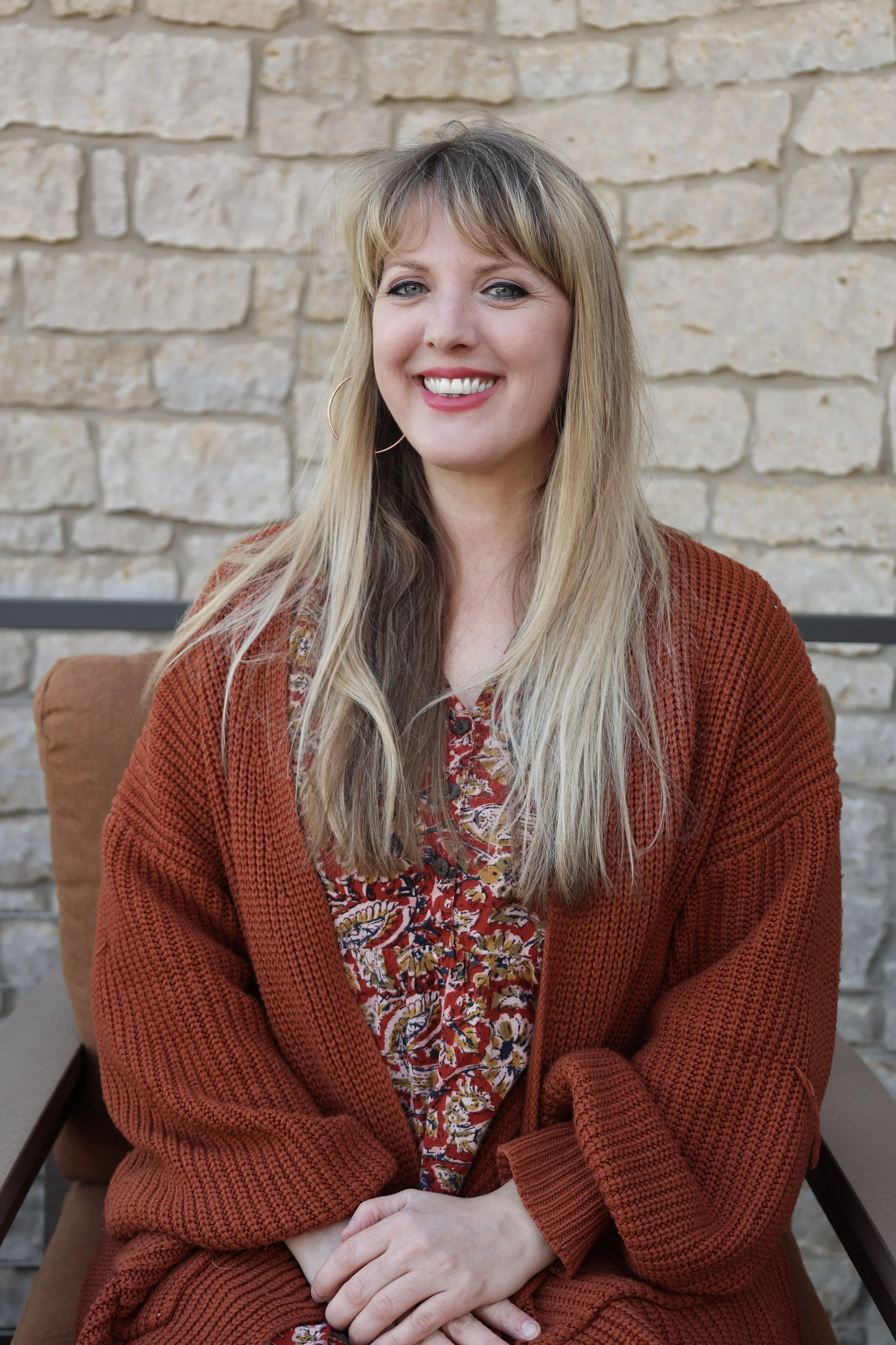 A woman with long blonde hair, smiling, seated indoors against a beige stone brick wall. She is wearing a floral dress and a rust-colored knitted cardigan.