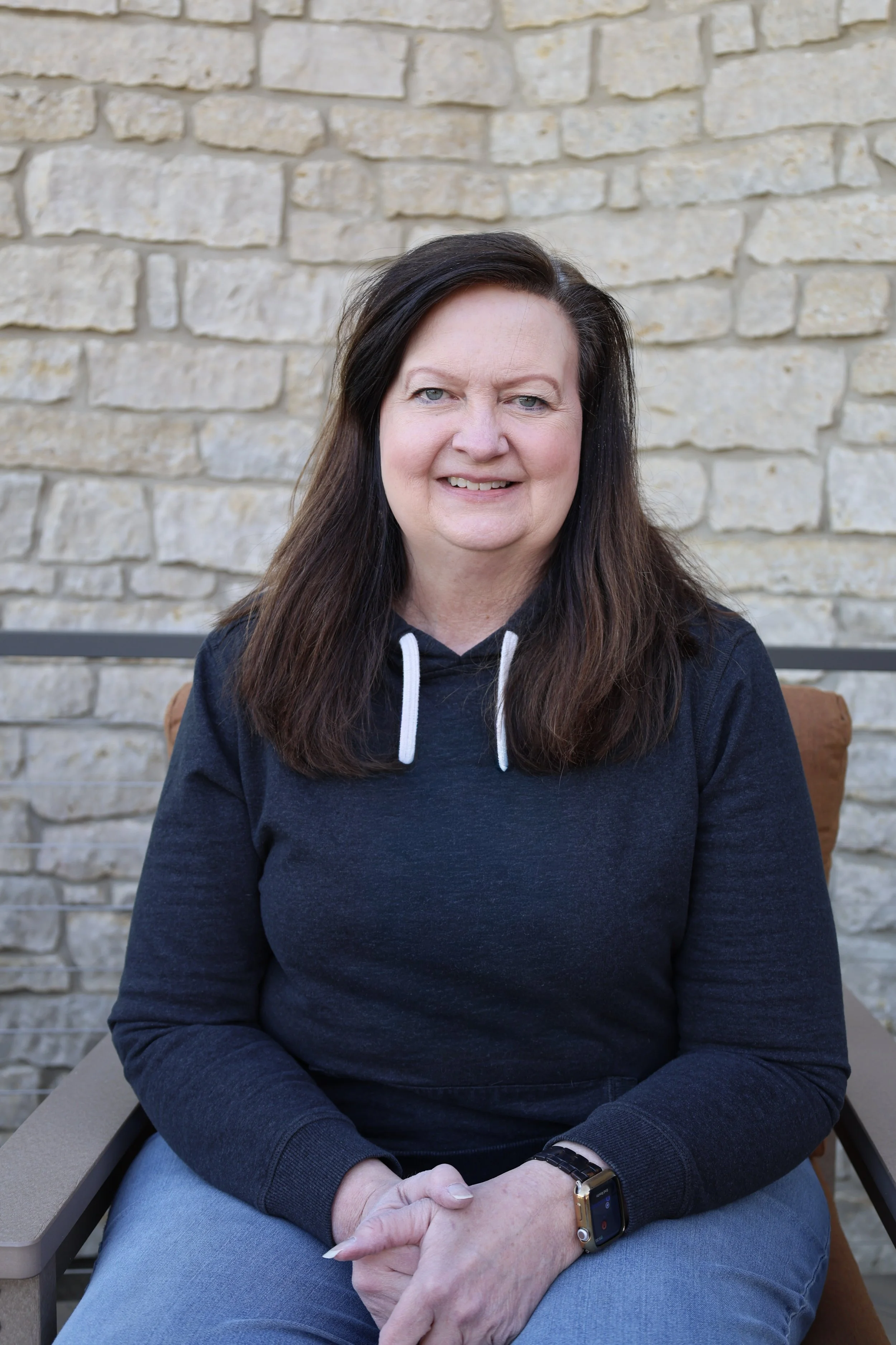 A woman with dark hair, blue eyes, and fair skin sitting outdoors in front of a stone wall. She is wearing a black hoodie with white drawstrings, grey pants, and a smartwatch on her left wrist. She is smiling slightly and looking at the camera.