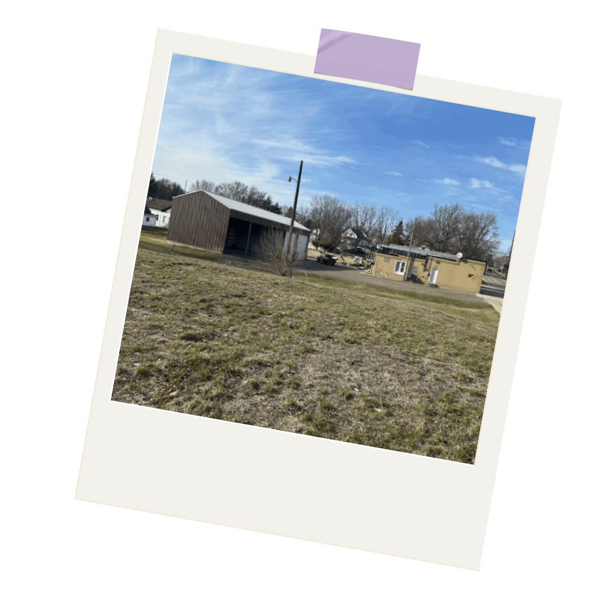 A small outdoor scene with a grassy area, a wooden building, a utility pole, and a yellow building with satellite dishes under a partly cloudy blue sky.