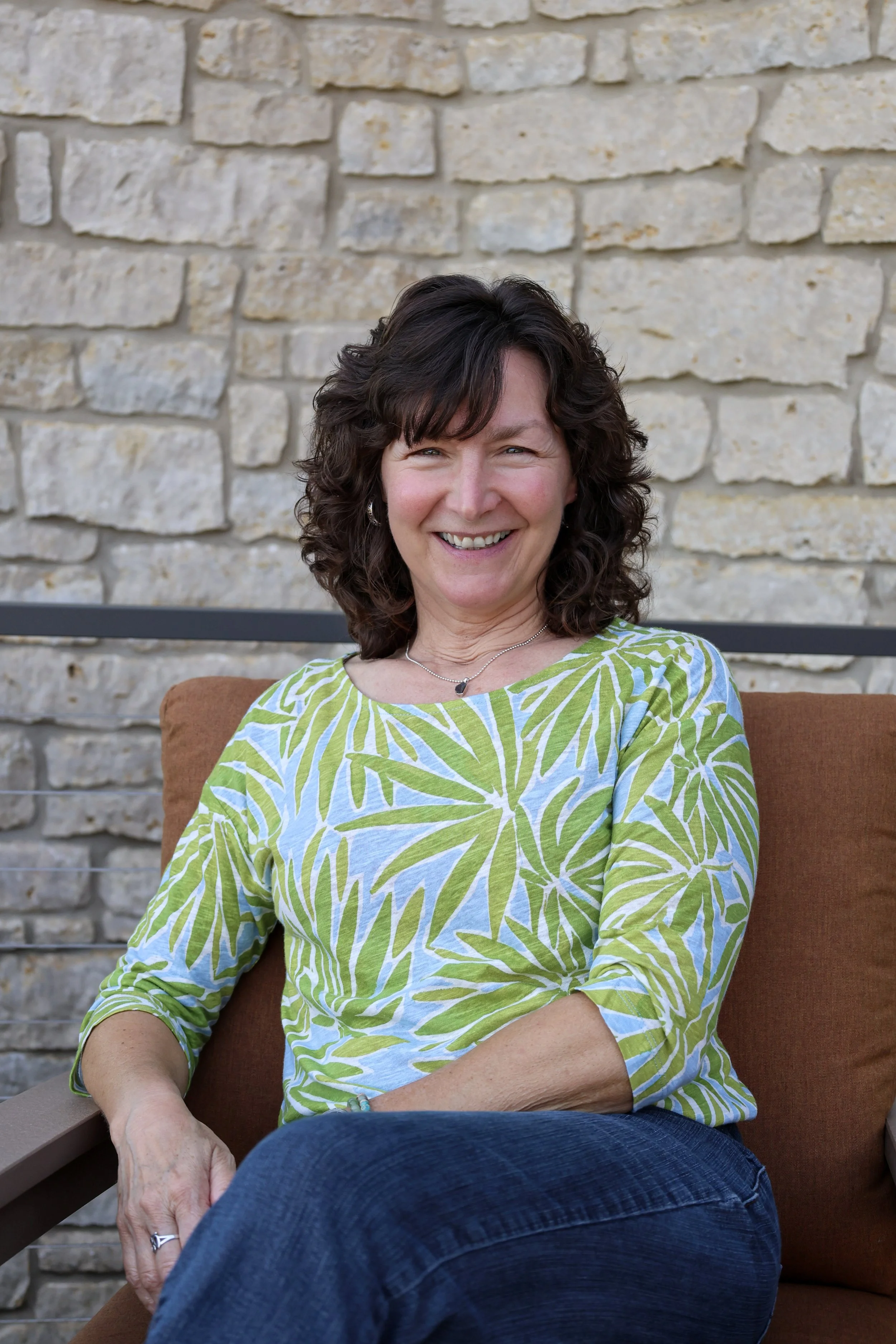 A woman with curly dark brown hair and light skin held back with bangs, smiling and wearing a green and white leaf-patterned shirt, sitting outdoors on a brown cushion with a beige brick wall behind her.
