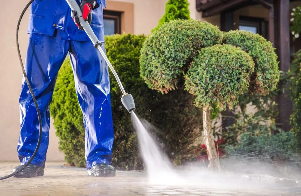 Person wearing blue waterproof pants using a pressure washer to clean a concrete surface outside a house, with bushes and trees in the background.
