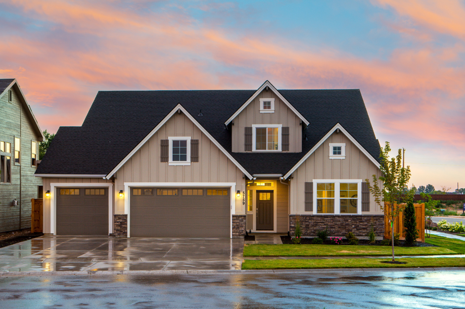 A modern house with beige siding, dark shutters, and a three-car garage, at sunset with a cloudy sky, wet driveway, and landscaped yard.