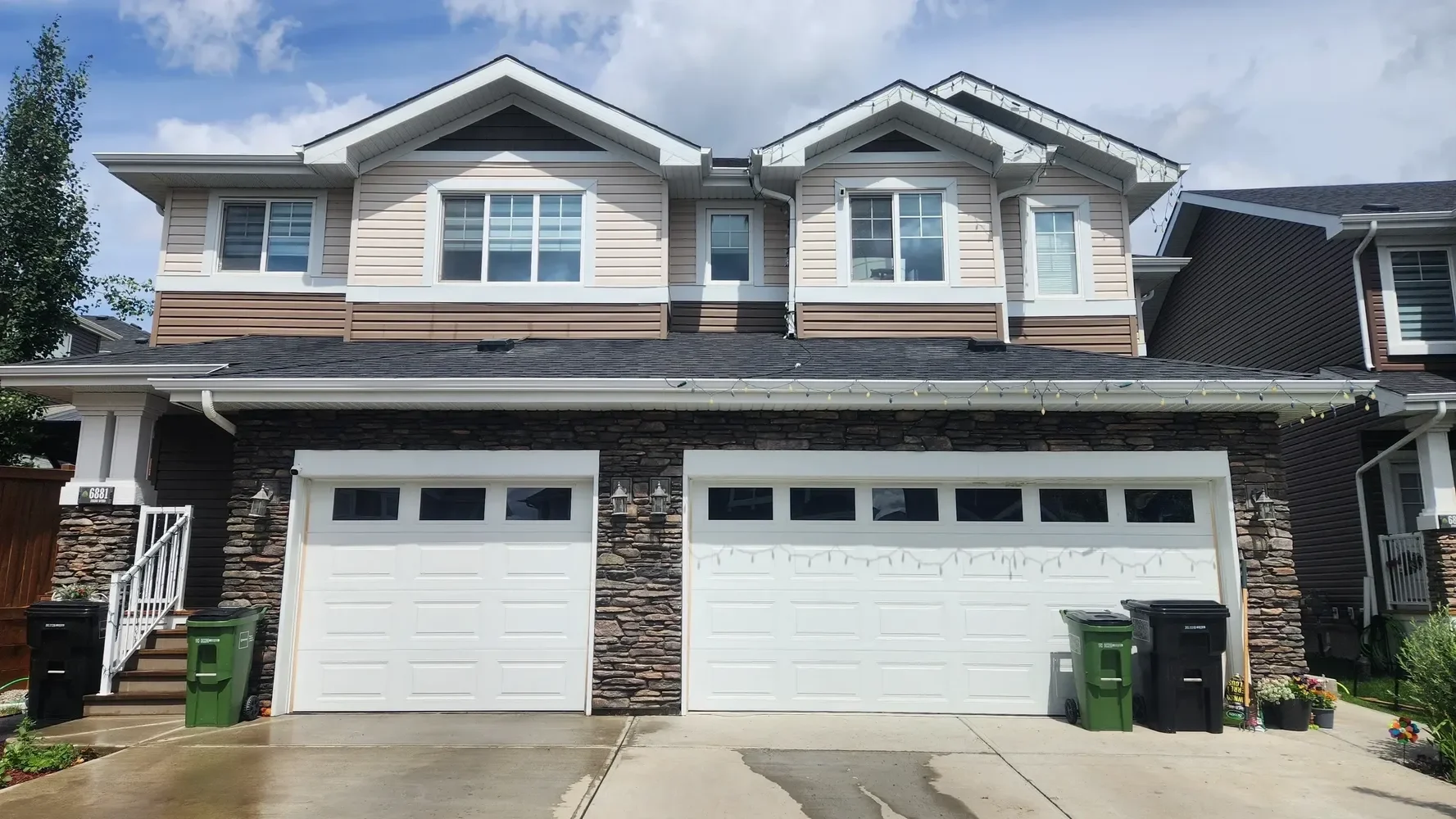 Modern two-story house with a two-car garage, stone facade, and tan siding. The house has multiple windows, some with white trim, and a dark shingled roof. There are green and black trash bins in front of the garage, and decorative plants and flowers near the entrance. String lights are hanging along the edge of the roof.