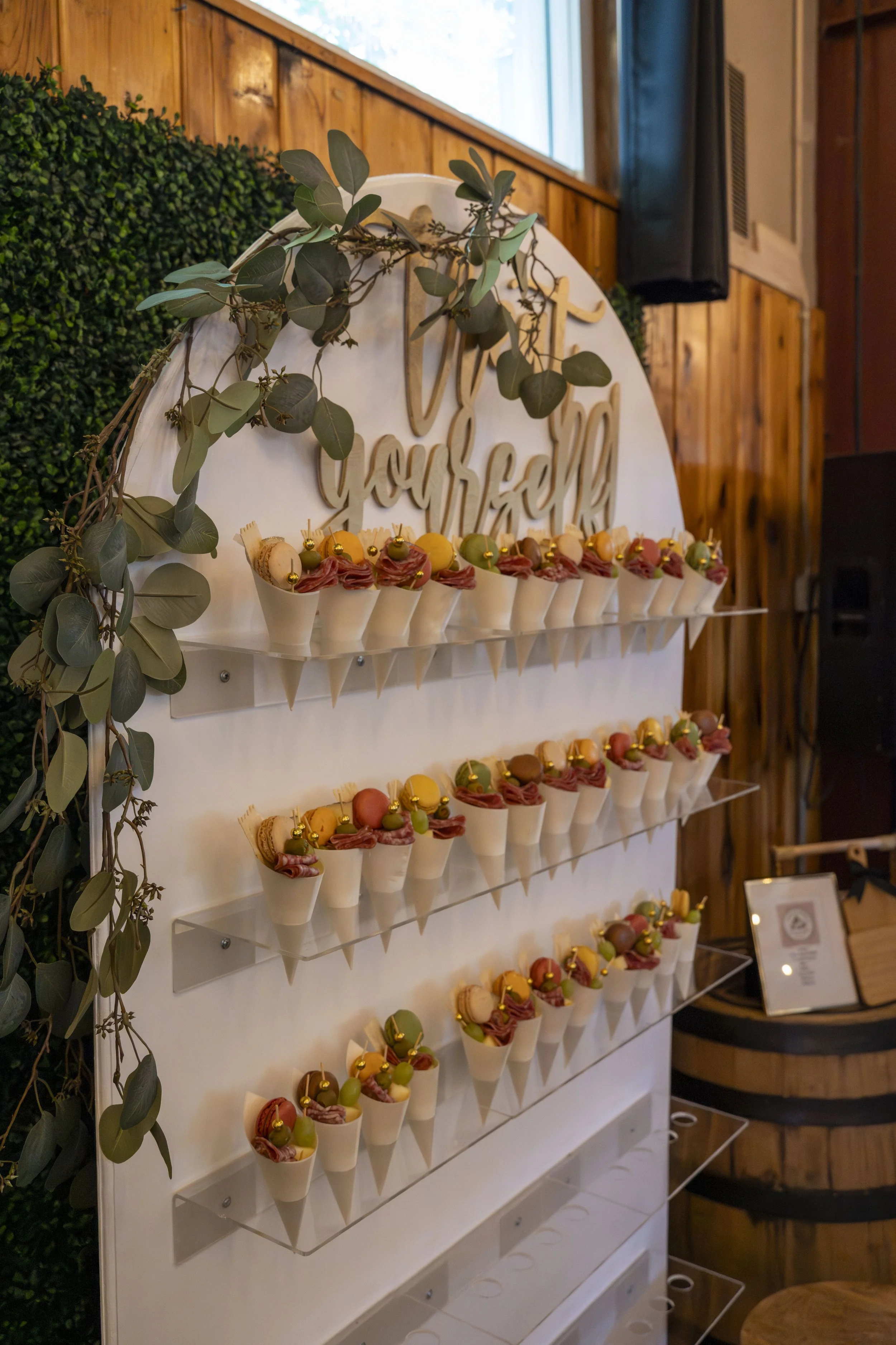 Display of colorful desserts with gold toppings on a white shelf, decorated with green leaves and a wooden 'You're' sign in a cozy indoor setting.