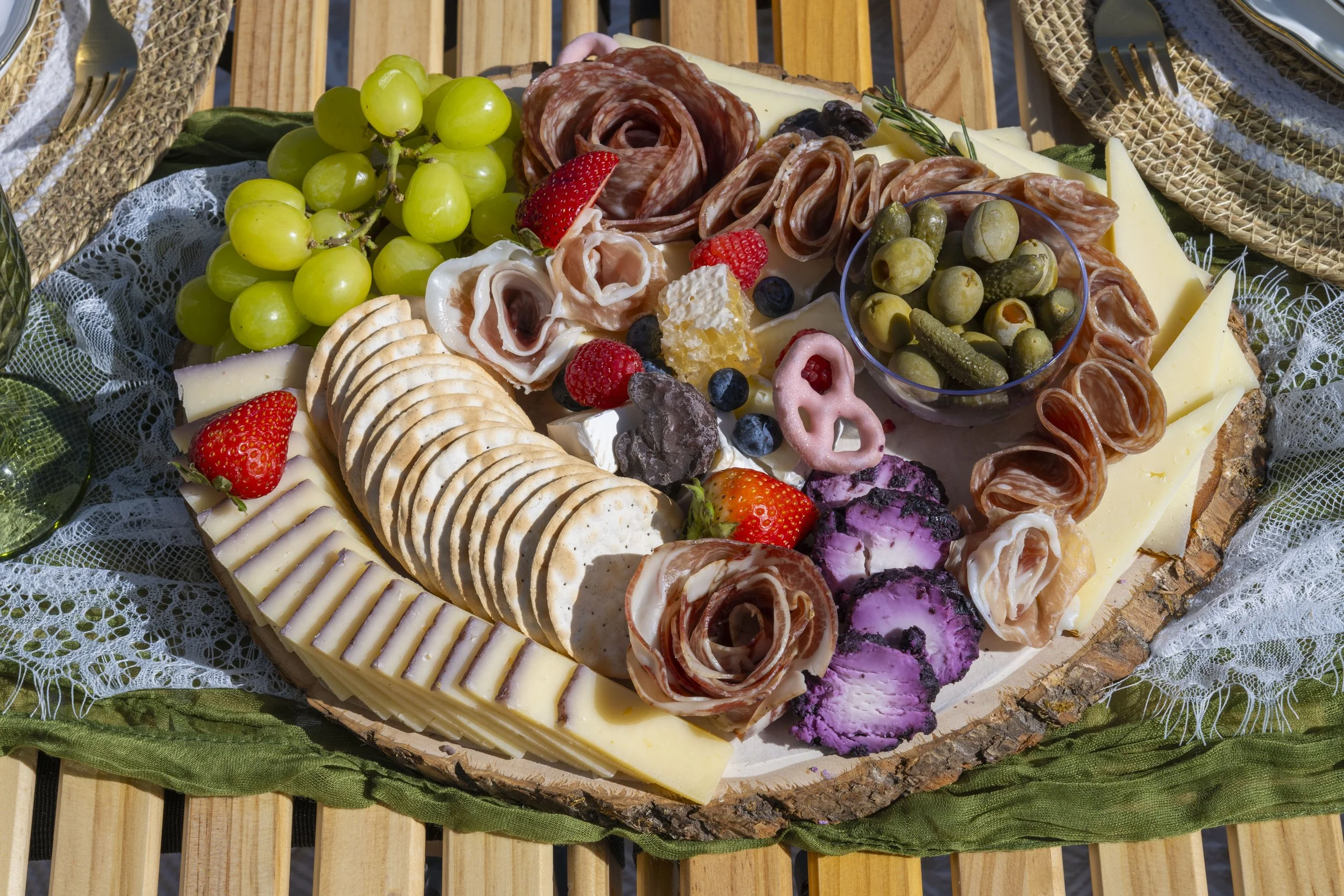 A cheese and charcuterie platter with grapes, strawberries, blueberries, olives, assorted cheeses, and cured meats on a wooden board with lace and napkins.
