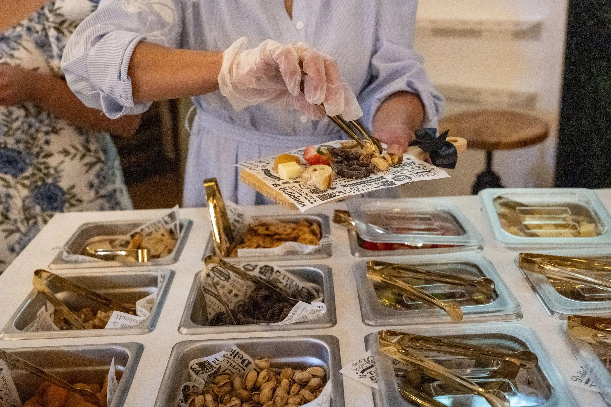 A person wearing a white shirt and gloves is serving themselves food from a buffet with various dishes in metal containers, including nuts, dried fruit, and other appetizers.