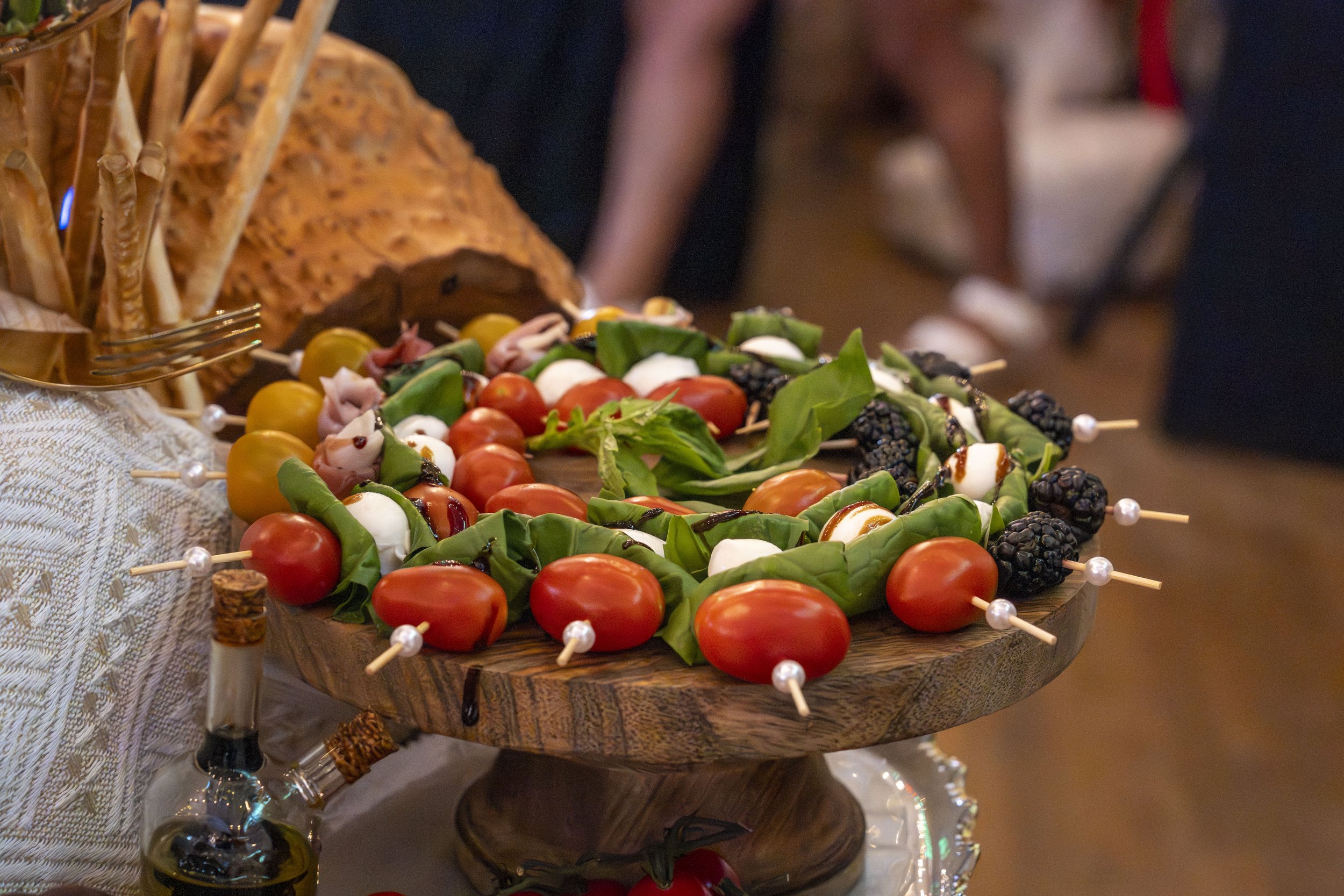 A wooden tray of fresh vegetable skewers with cherry tomatoes, mozzarella, basil, and blackberries, surrounded by crackers, on a table at a social gathering.