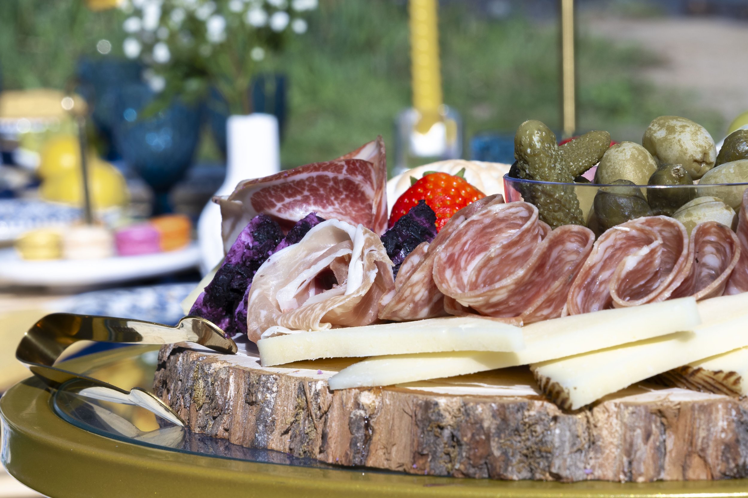 Assorted sliced meats, cheese, strawberries, and pickles on a wooden serving platter with a gold-colored tray underneath, outdoors with blurred greenery and tableware in the background.