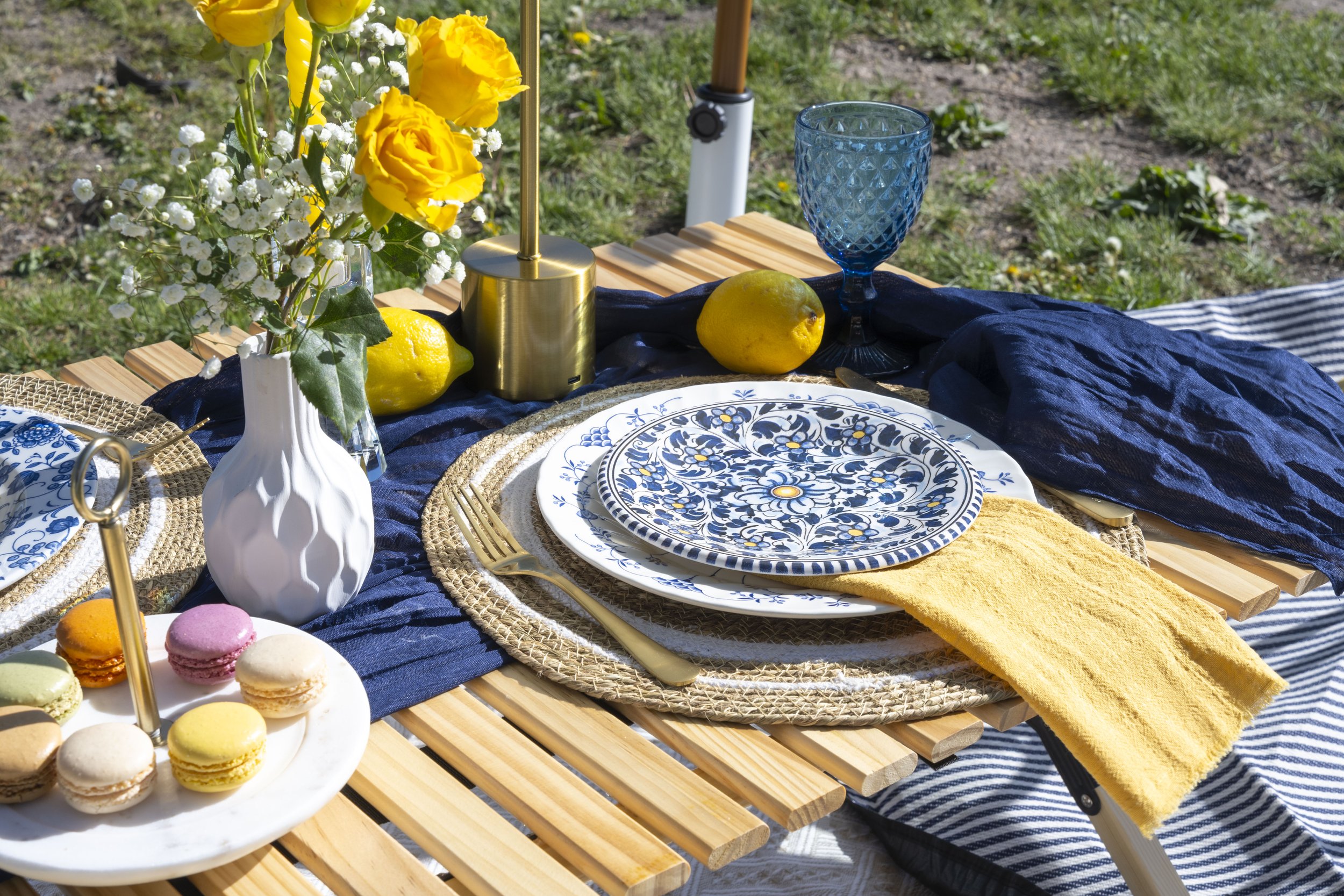A picnic table with a blue tablecloth set with decorative plates, a bouquet of yellow flowers, two lemons, a blue goblet, a white pitcher, and a plate of colorful macarons, outdoors on grass.