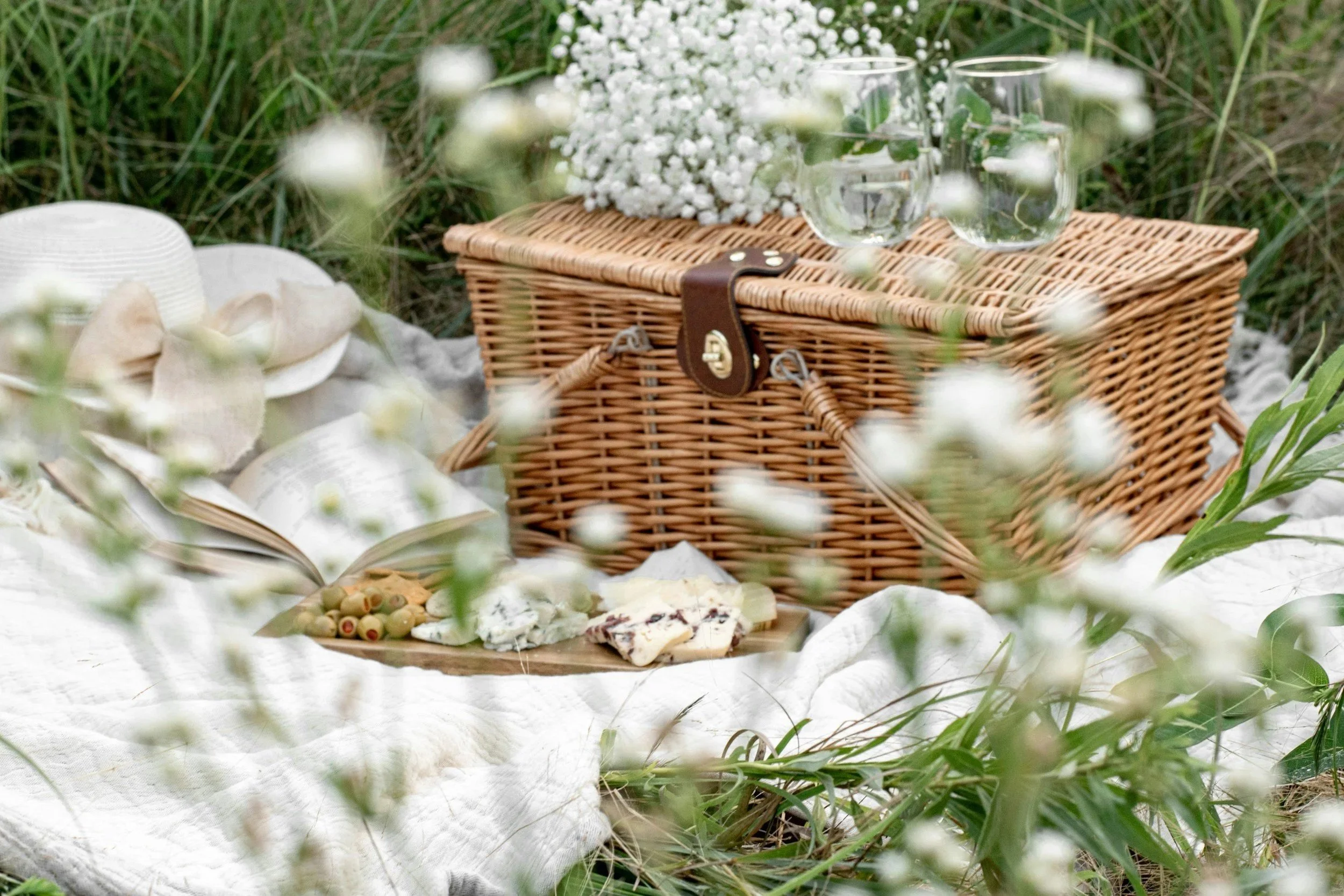 A picnic setup featuring a wicker picnic basket with a leather strap, surrounded by white flowers and greenery, with a white hat and open notebook on a white cloth.