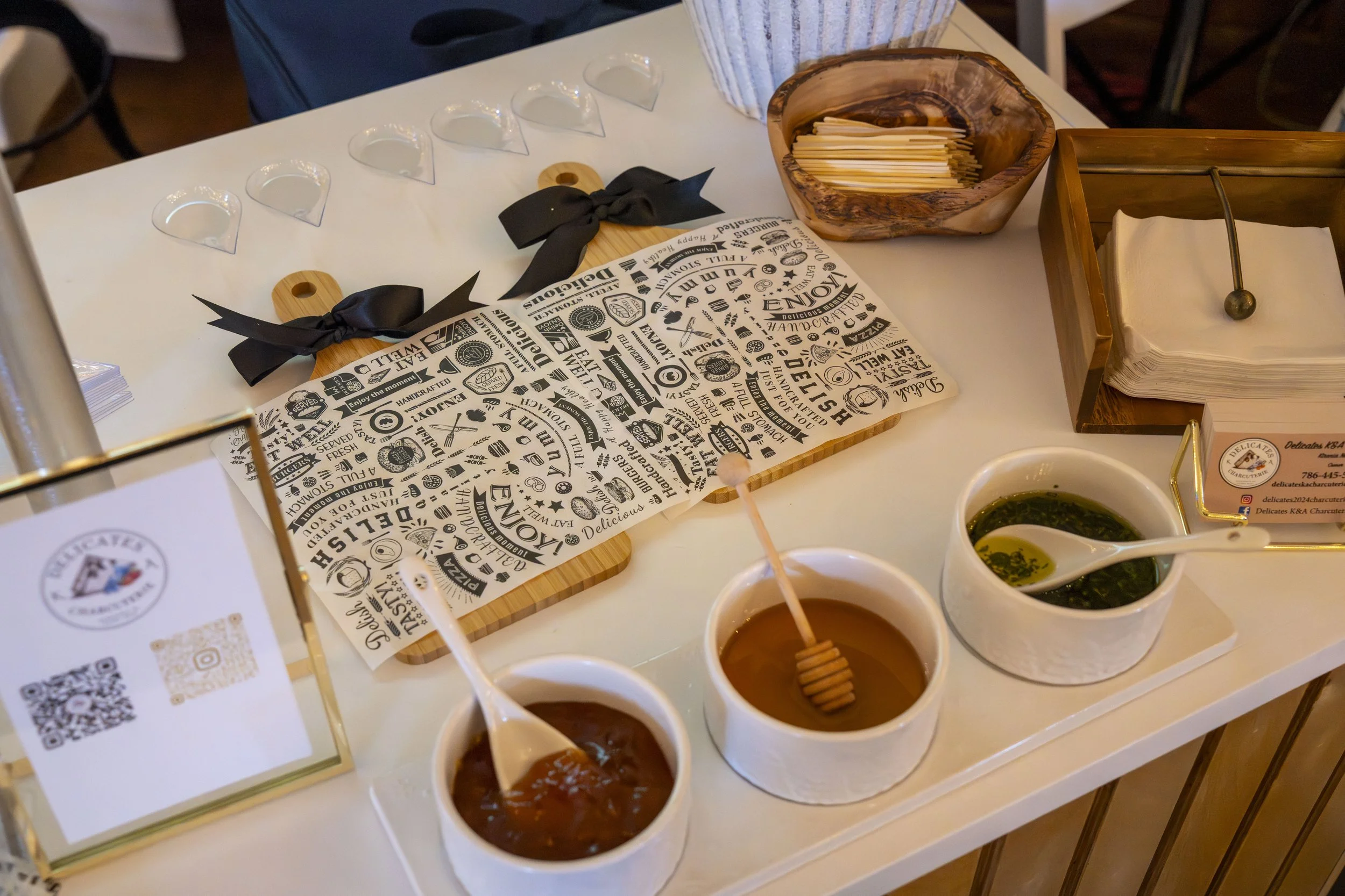 Assorted sauces in small bowls with honey, placed on a white table next to a chalkboard menu with pictures and text, a wooden basket with utensils, and a cardboard box containing napkins, in a restaurant setting.