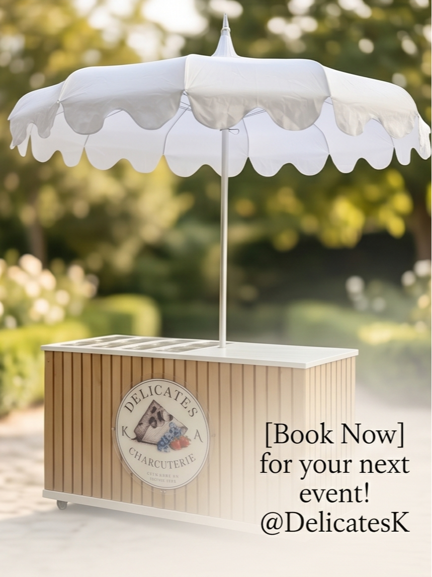 A mobile food cart with a white scalloped umbrella, wooden slat design, and a circular logo that says "Delicates Charcuterie" with images of cheese, grapes, and a strawberry.