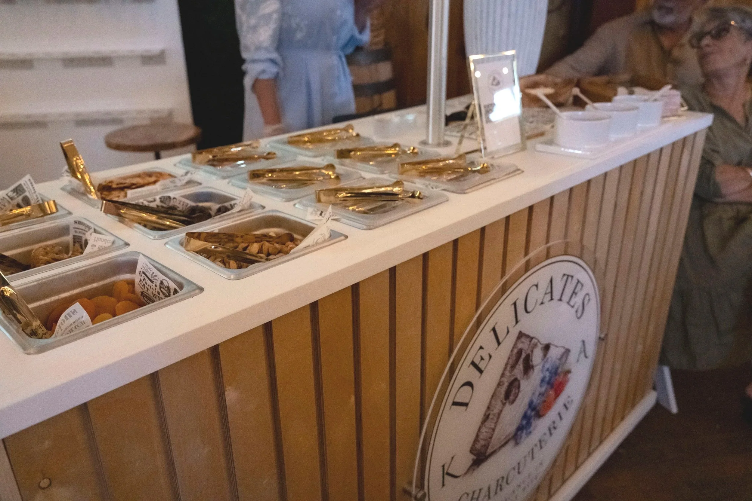 A dessert bar with various toppings in small containers, including nuts and candies, on a white counter with wooden paneling. There are people in the background.