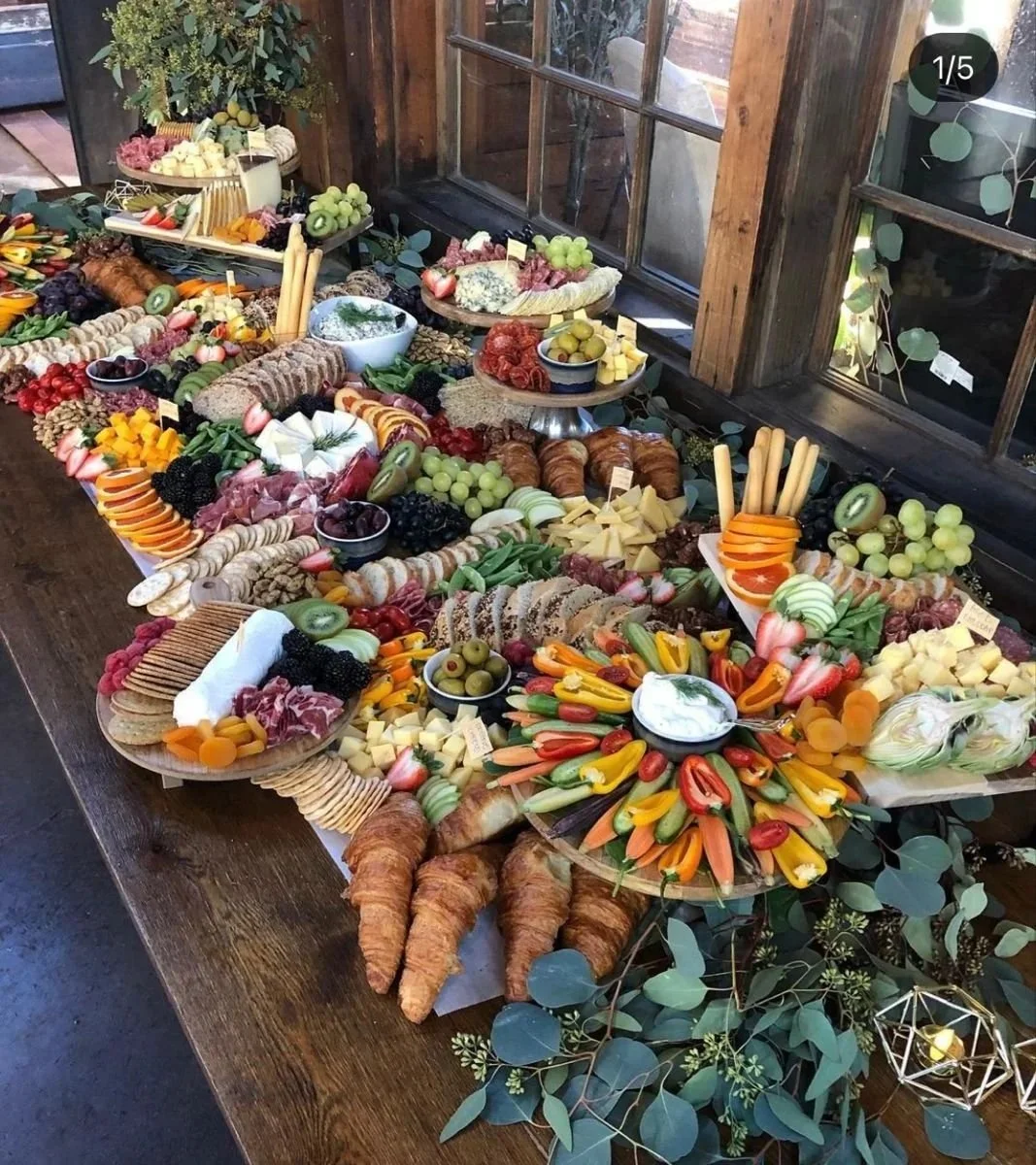 A table filled with a variety of fruits, vegetables, cheeses, crackers, bread, and charcuterie, set in a rustic setting near a window with greenery and decorative elements.
