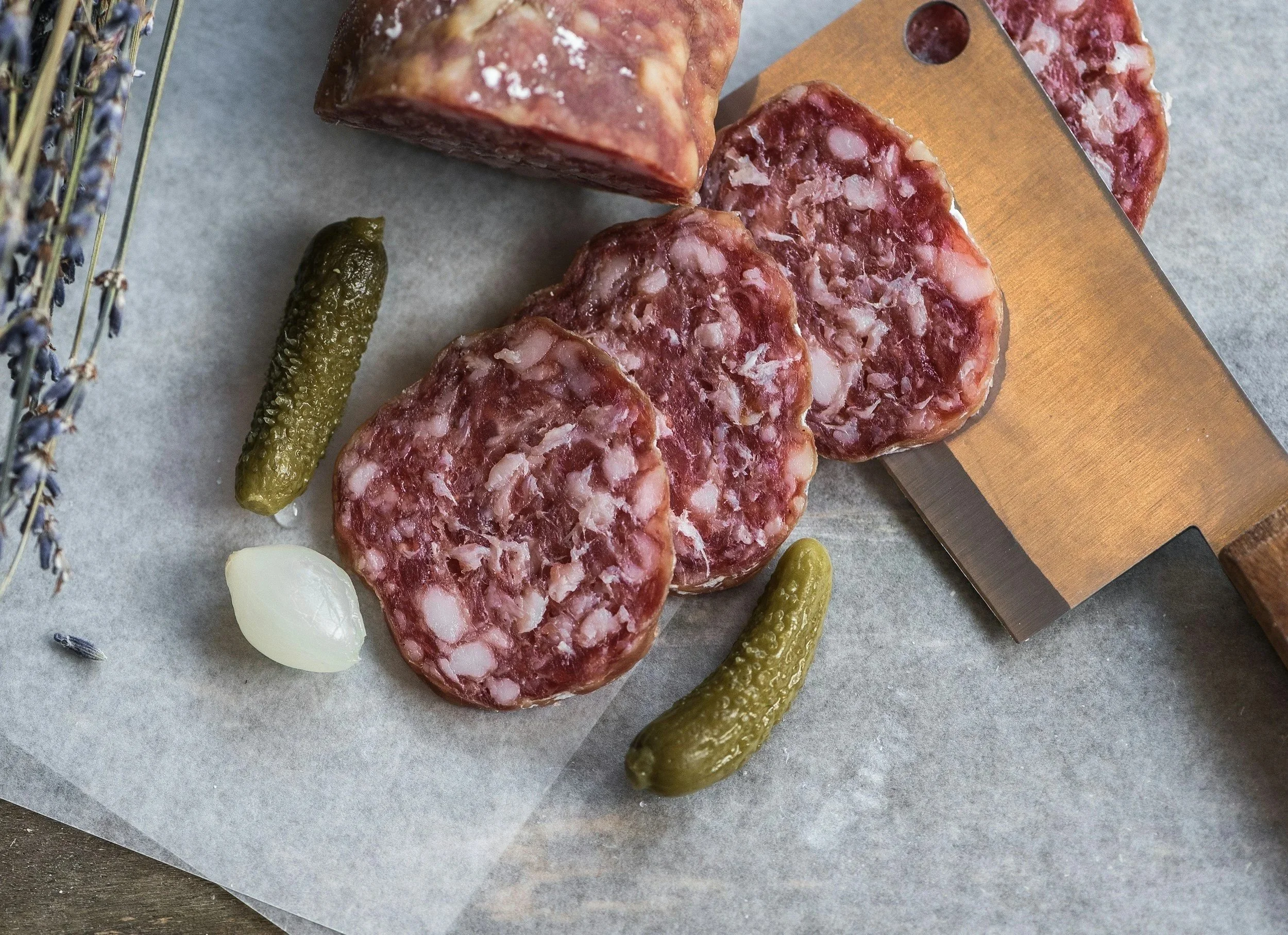 Slices of cured sausage with a marbled appearance, placed on parchment paper, alongside small pickles, garlic, lavender sprigs, and a large meat slicing knife.