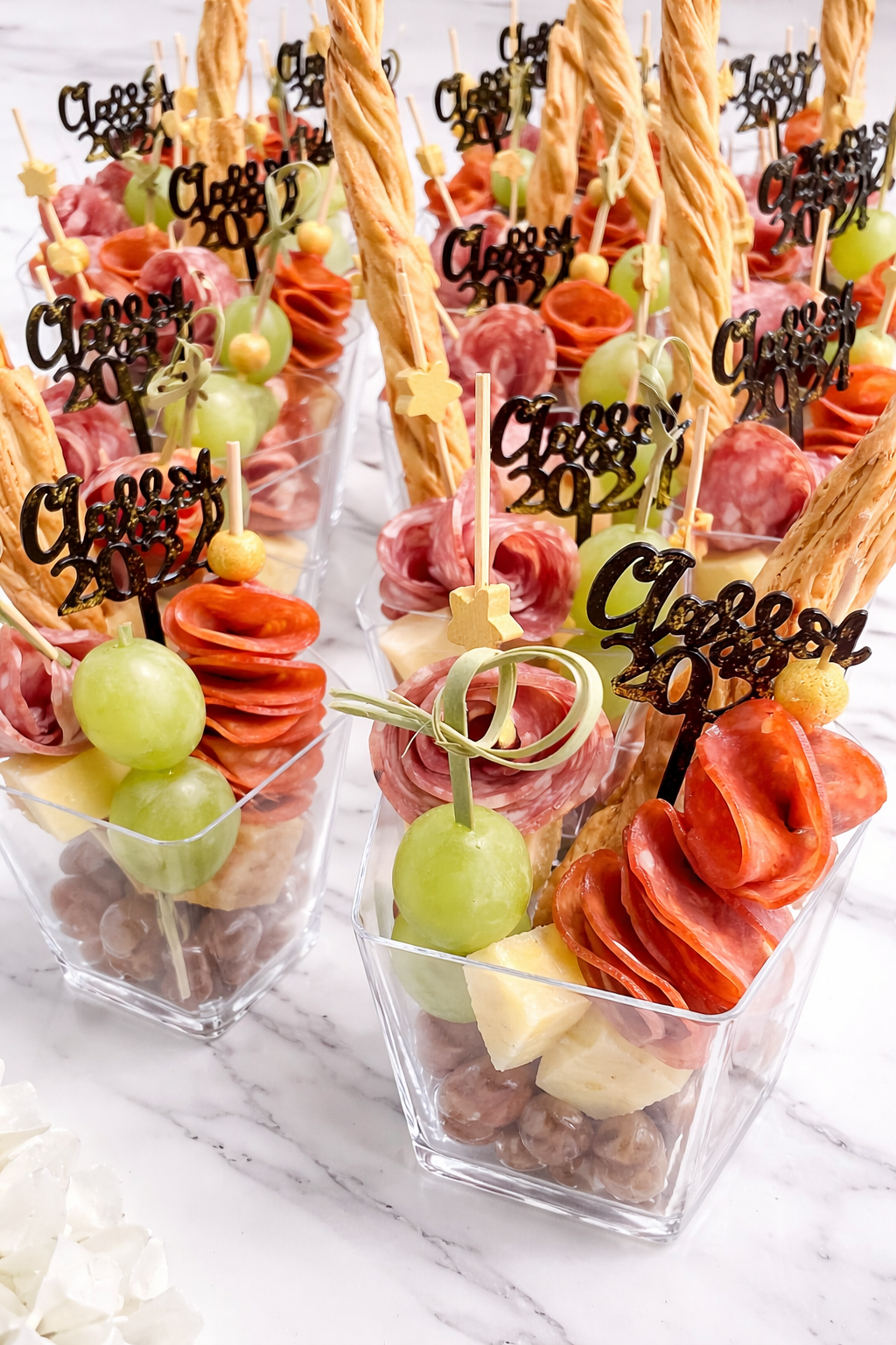 Party appetizer cups filled with sliced meats, cheese, green grapes, and nuts, each with a decorative 'Celebrate 2024' topper and a breadstick.