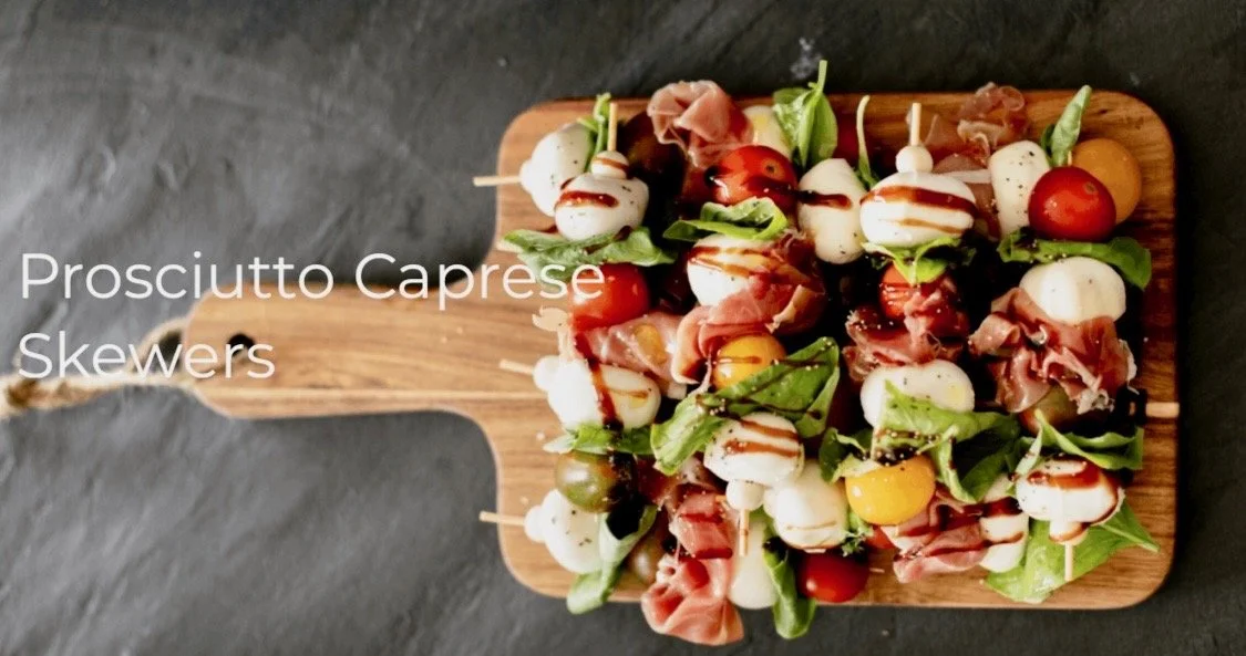 Prosciutto Caprese skewers on a wooden serving board with cherry tomatoes, mozzarella balls, basil leaves, and balsamic glaze.