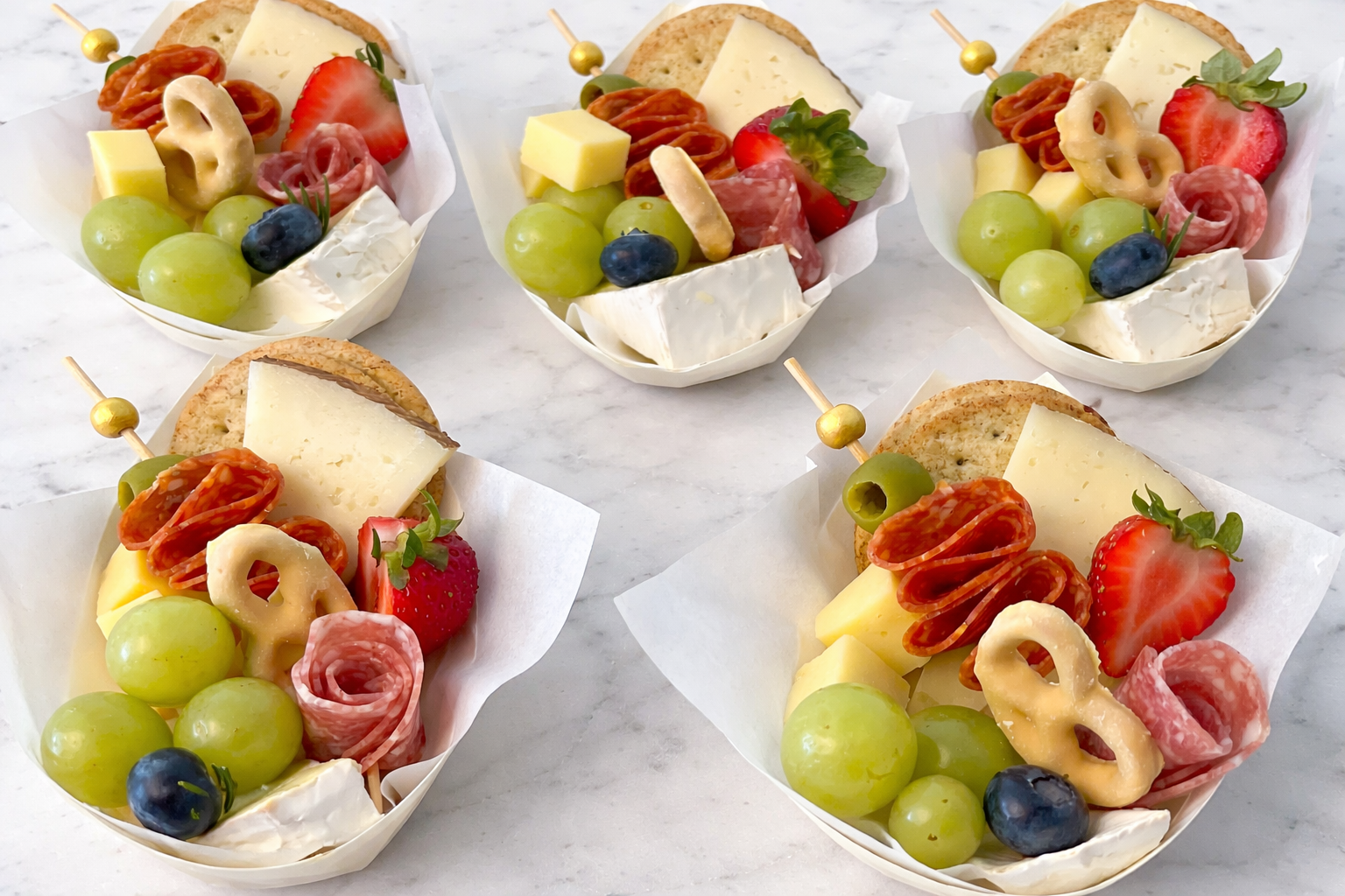 Five small cheese and fruit platter snacks on a white marble surface, each containing green grapes, blueberries, strawberries, slices of cheese, slices of cured meat, a cracker, and a small cheese wedge, with a decorative toothpick.