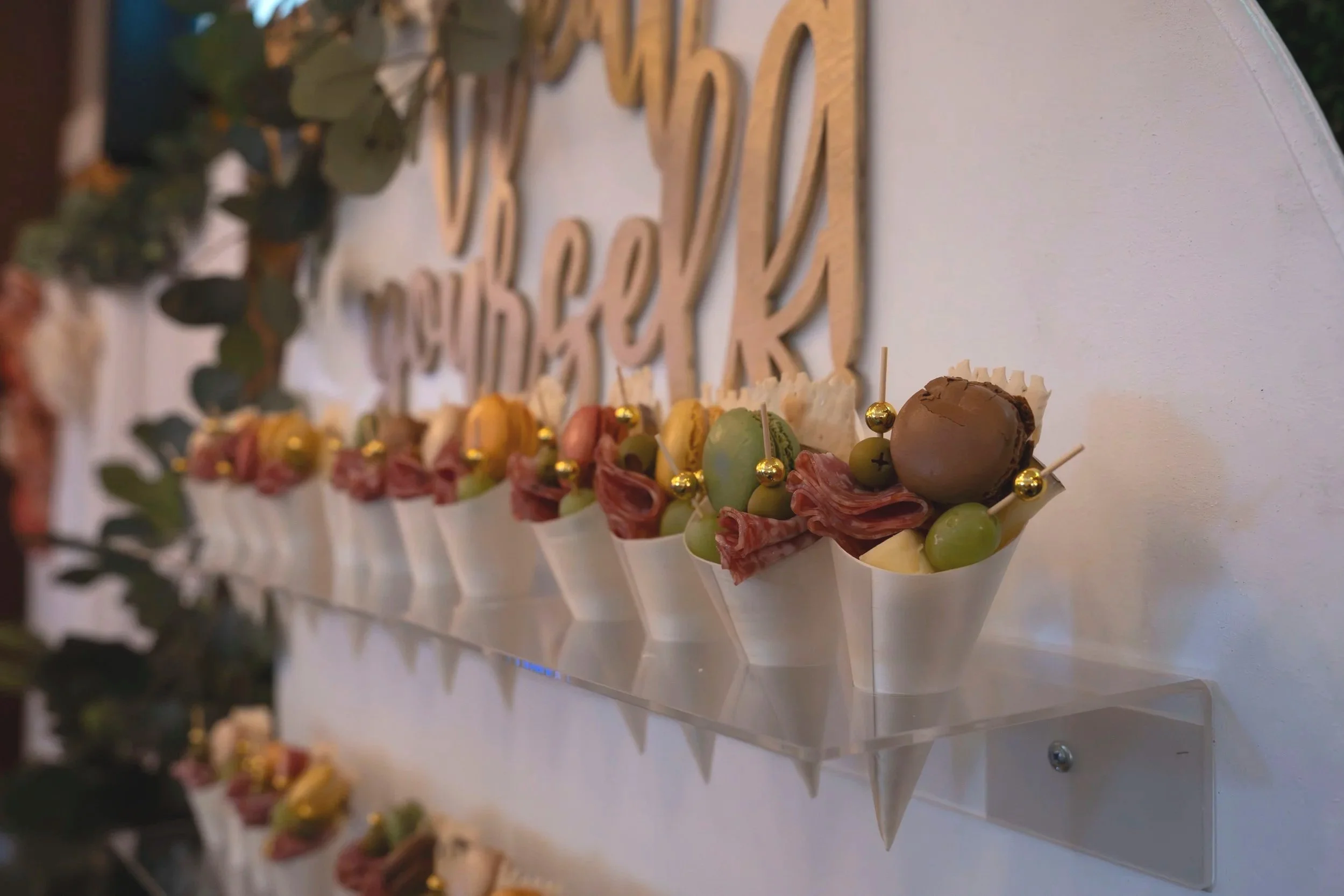 Close-up of appetizer cups filled with various meats, cheeses, green grapes, and olives, decorated with gold-colored beads, with a wooden sign with the word 'WEDDING' in the background.