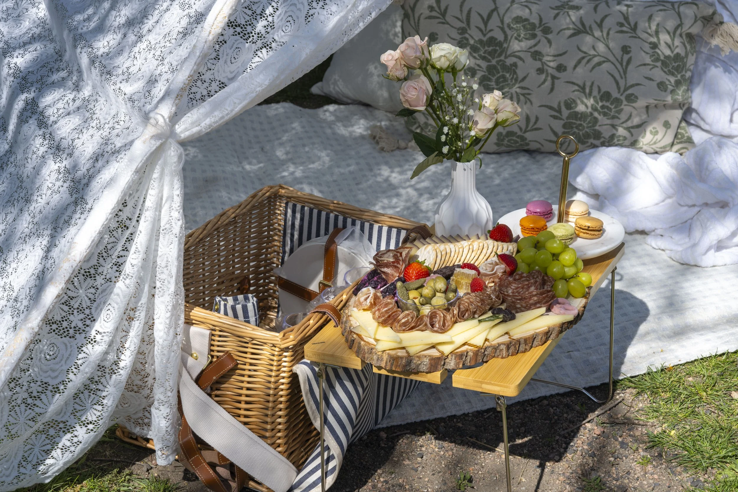 A picnic setup with a wooden serving board filled with cheese, meats, strawberries, grapes, and macarons. A white vase with pink and white roses is on a small table. The table is covered with a white and blue striped cloth. Behind the setup, there are white lace curtains and cushions with floral patterns. It appears to be outdoors on grass.