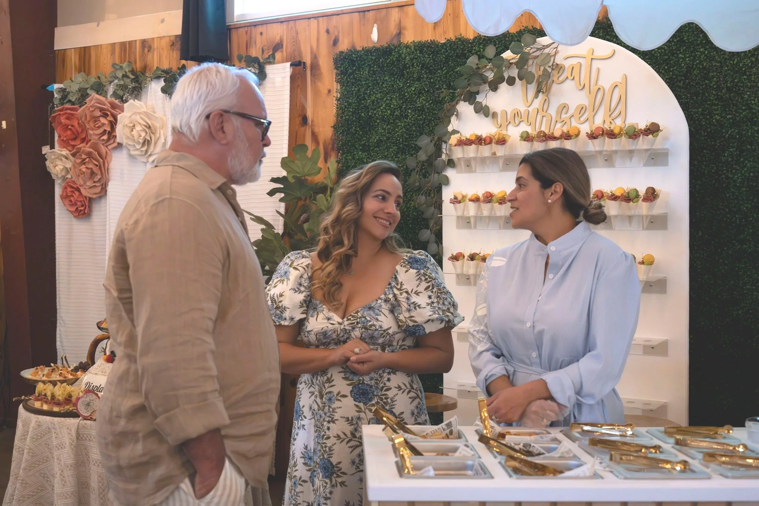 Three women and an older man engaged in conversation at a dessert table during a celebration event with a decorative backdrop saying 'treat yourself'.