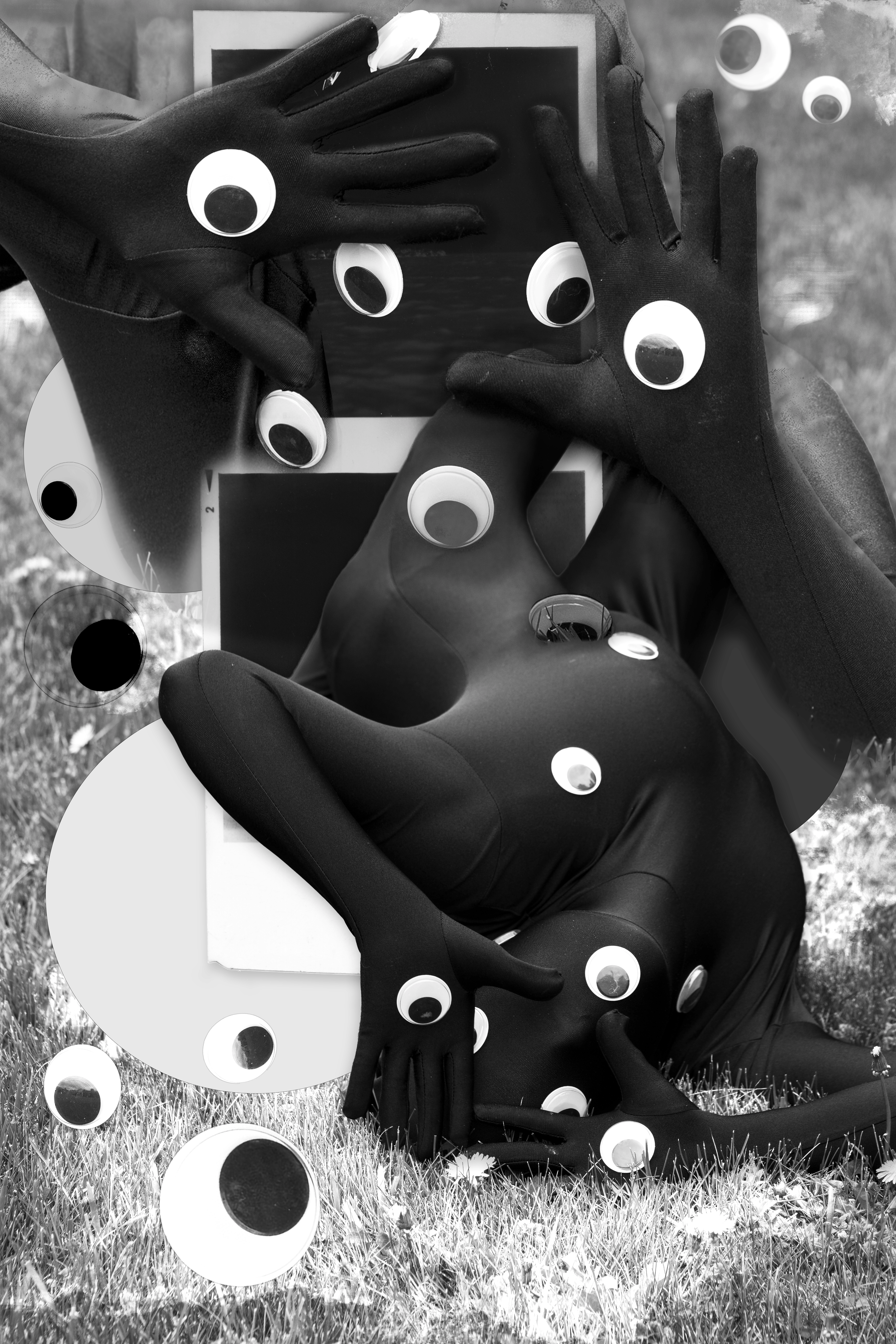 Black and white photo of a person in a stretchy bodysuit lying on grass, holding a tablet. The person and the tablet are decorated with large paper eyes. The photo has a surreal, playful style.