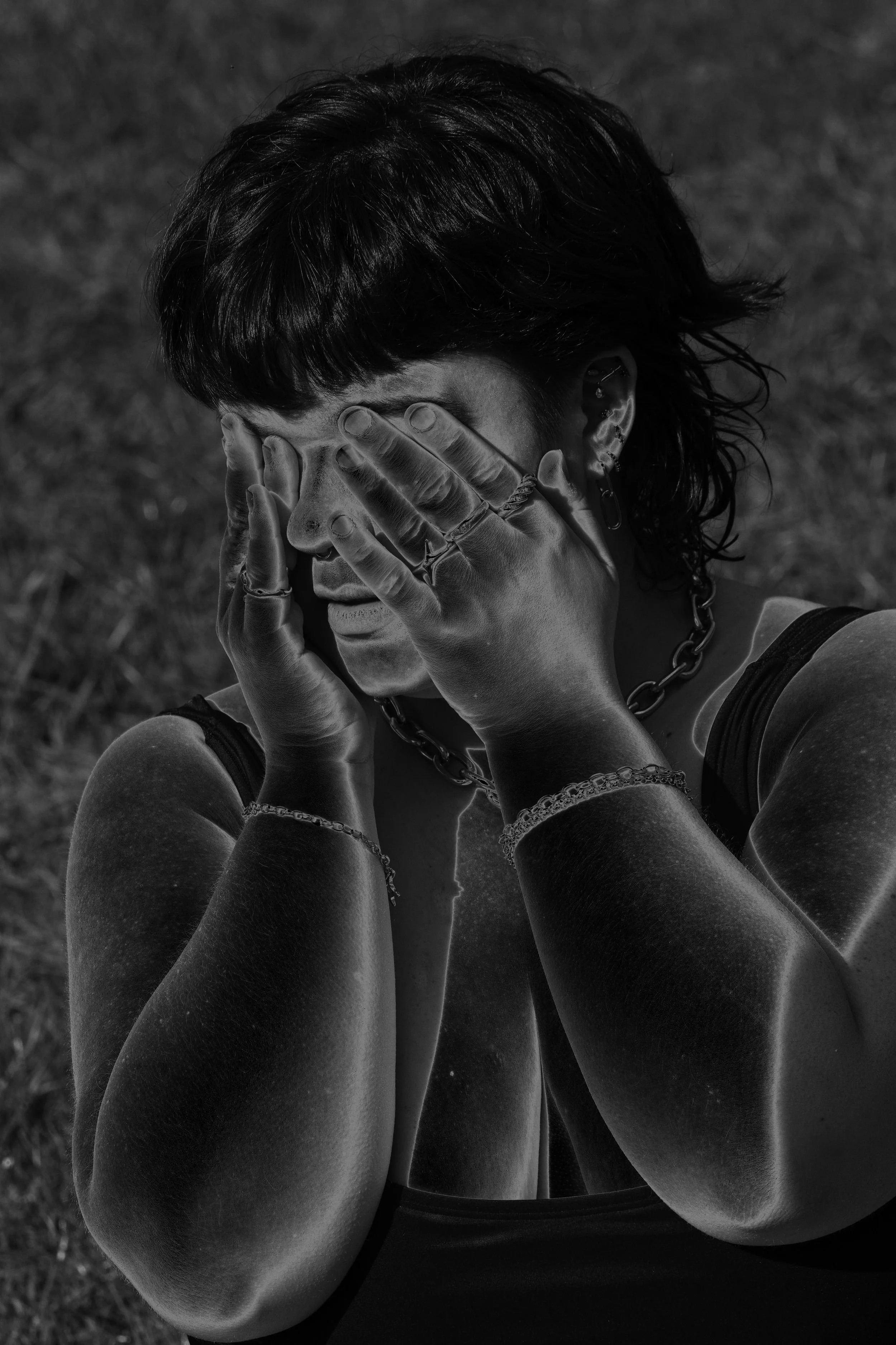 Black and white photo of a woman with short hair covering her face with her hands, wearing necklaces and bracelets, outdoors on grass.