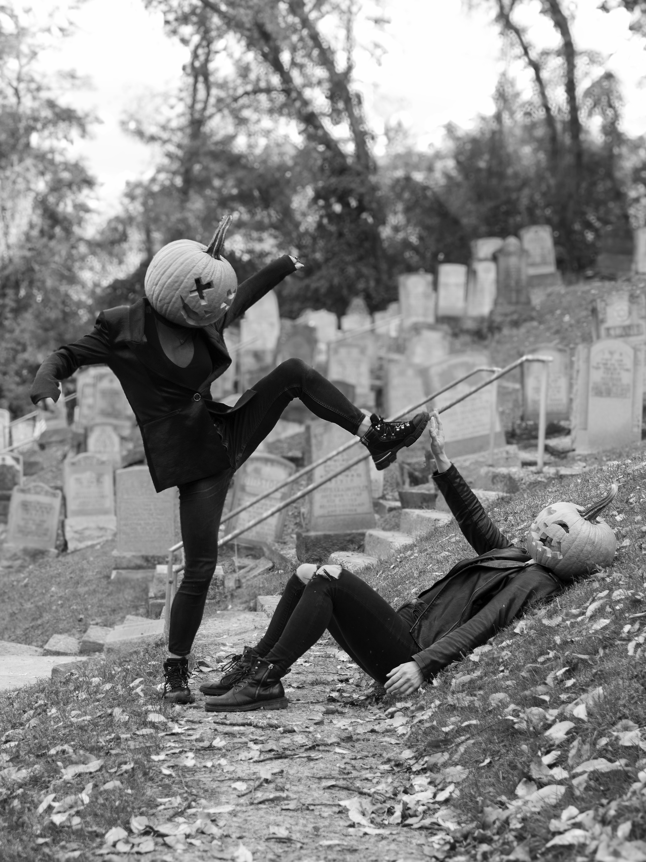Two people with pumpkin head masks posing as if fighting on a cemetery path, one lying on the ground and the other standing and punching, in a spooky Halloween scene.