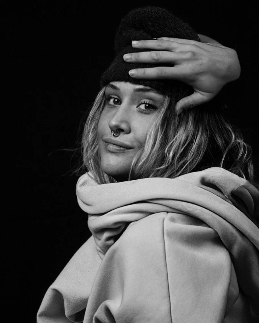 A young woman with wavy hair wearing a beanie, a septum piercing, a hoodie, and holding her hand on her forehead, looking at the camera with a slight smile in a black and white portrait.