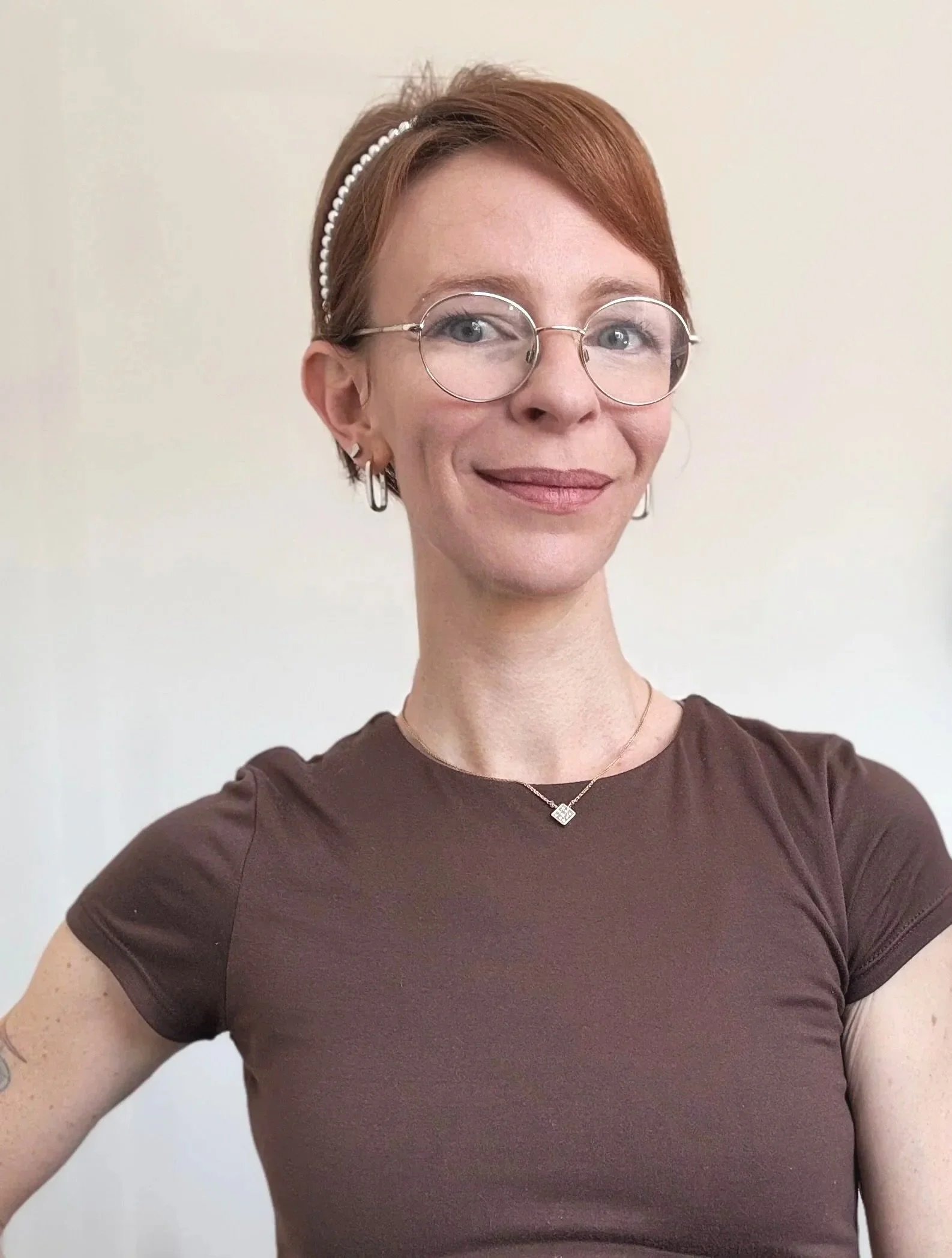 A woman with short red hair wearing glasses, a pearl hairband, earrings, a necklace with a pendant, and a brown shirt, standing against a plain white background.