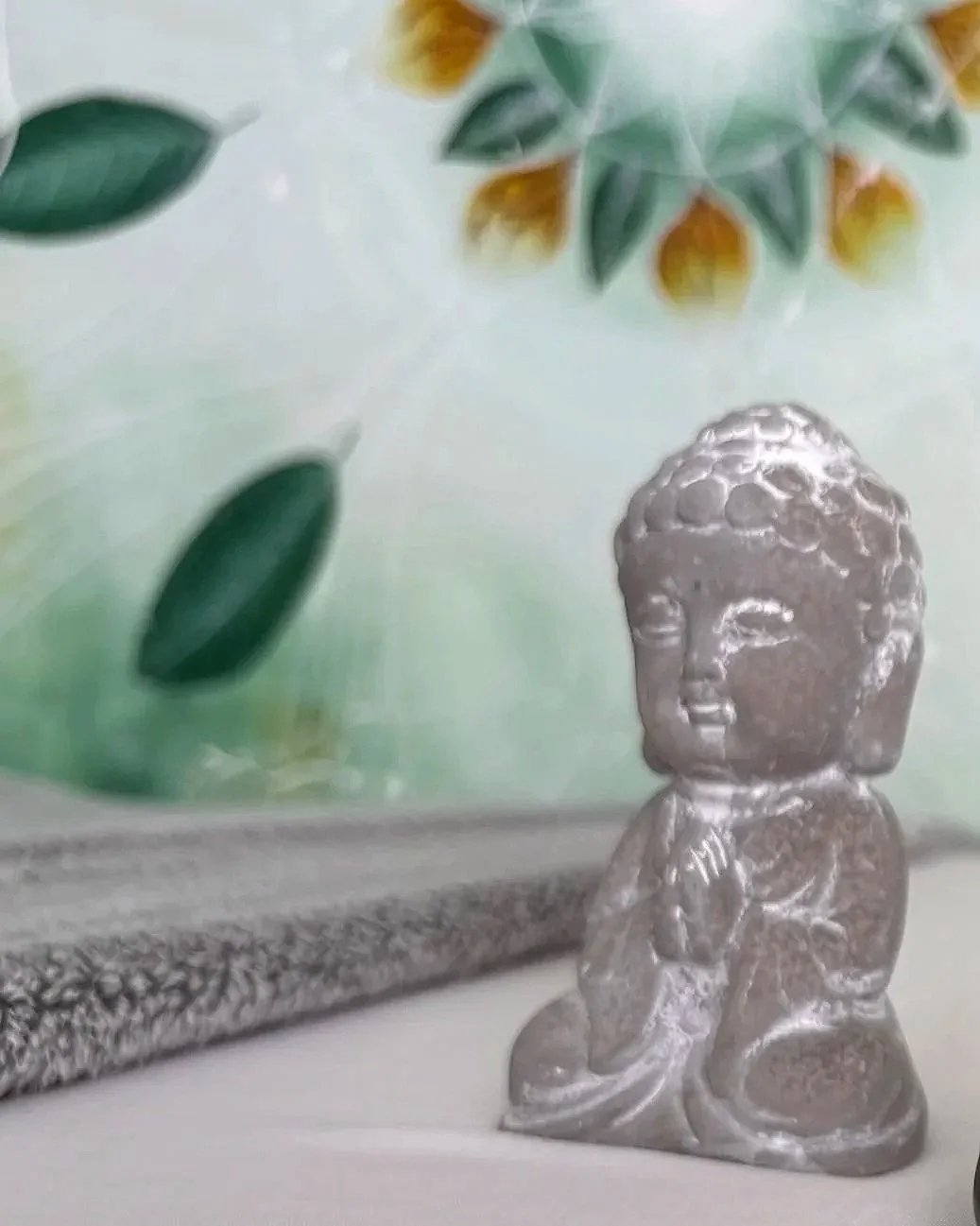 A small silver Buddha figure with clasped hands in a prayer position, placed on a flat surface, with decorative wall art featuring green leaves and yellow accents in the background.