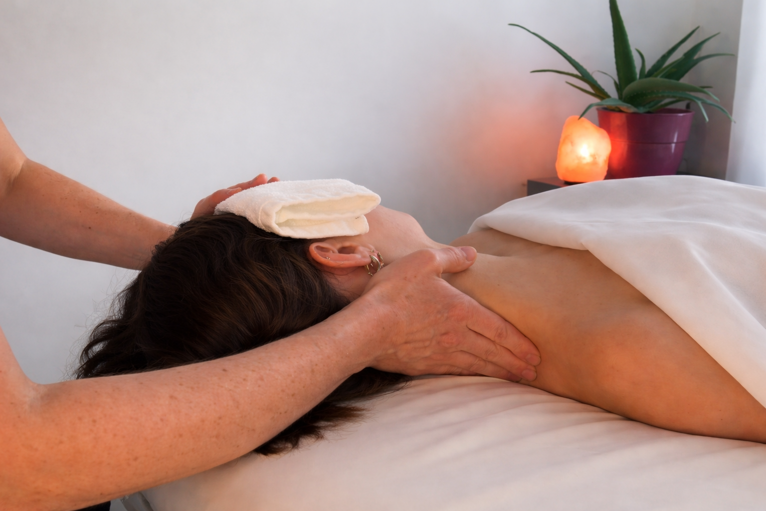Person receiving a massage on back in a massage room with a salt lamp and a potted plant on a nightstand in the background.