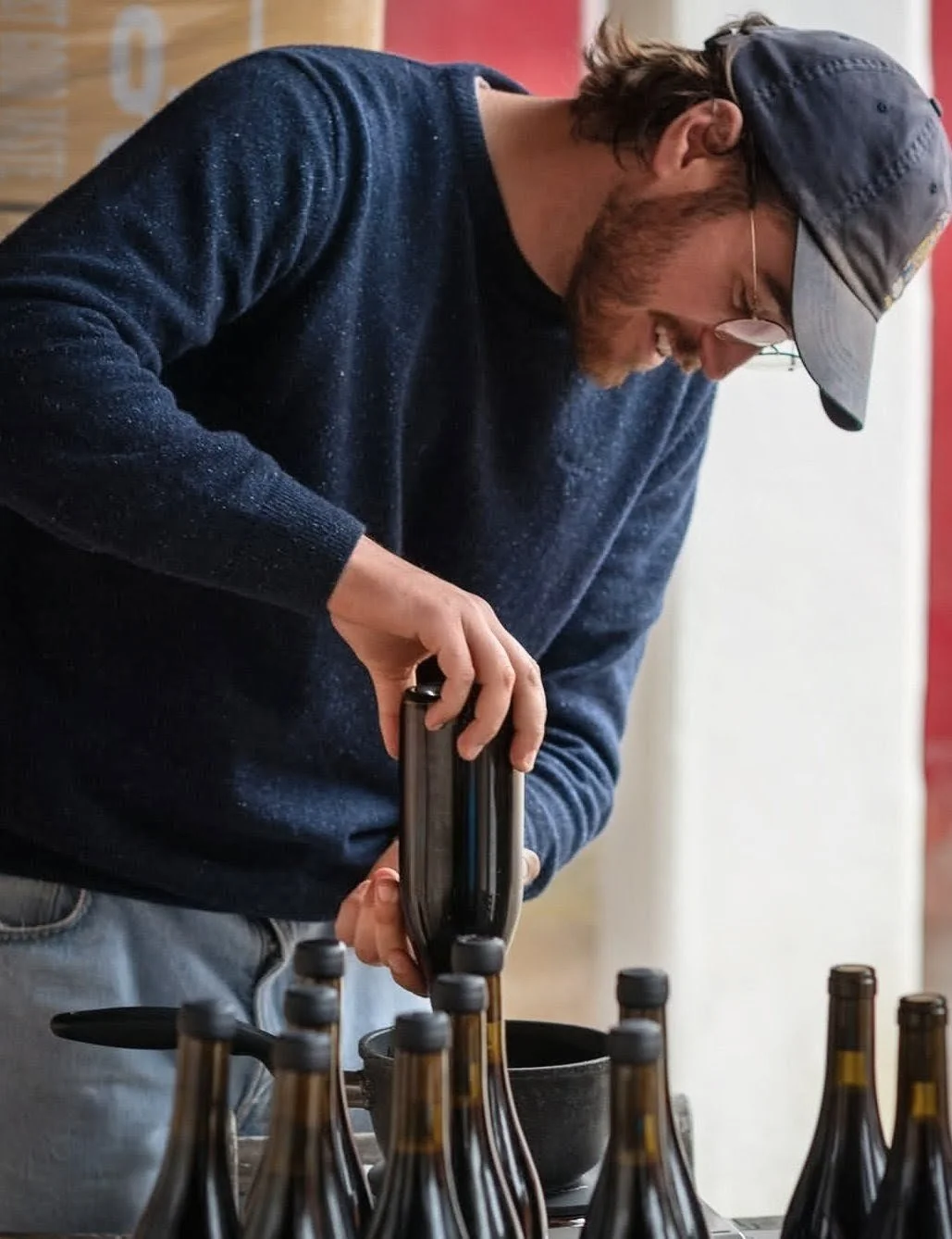 A man wearing a gray baseball cap, glasses, and a navy blue sweater with specks, is pouring beer from a tap into a glass at a brewery or bar. Several beer bottles are on the table in front of him.