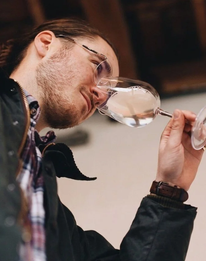 A man with glasses and a beard holding a wine glass and smelling its contents.