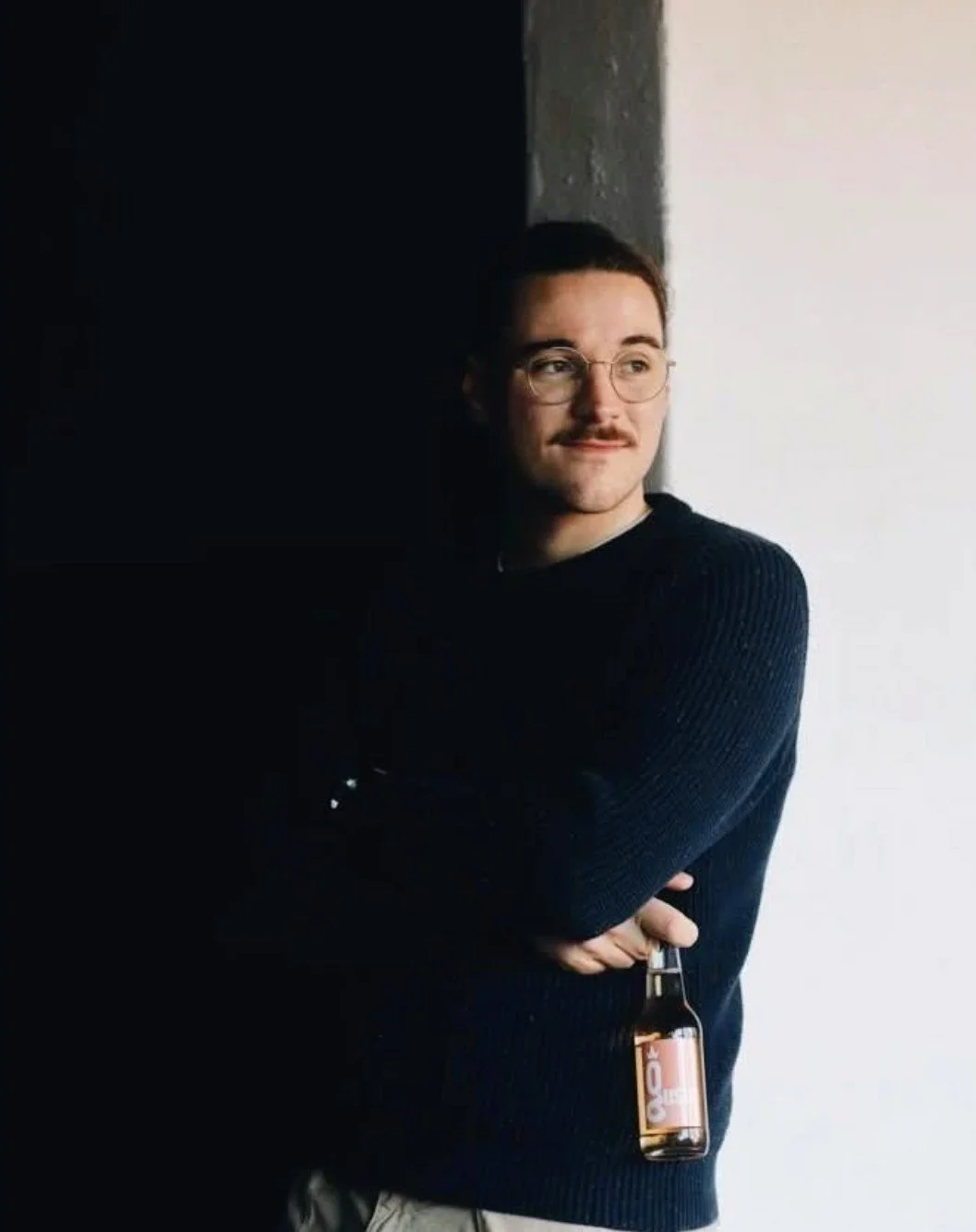 A young man with glasses and a mustache stands against a wall, holding a bottle of beer, with a contemplative expression.