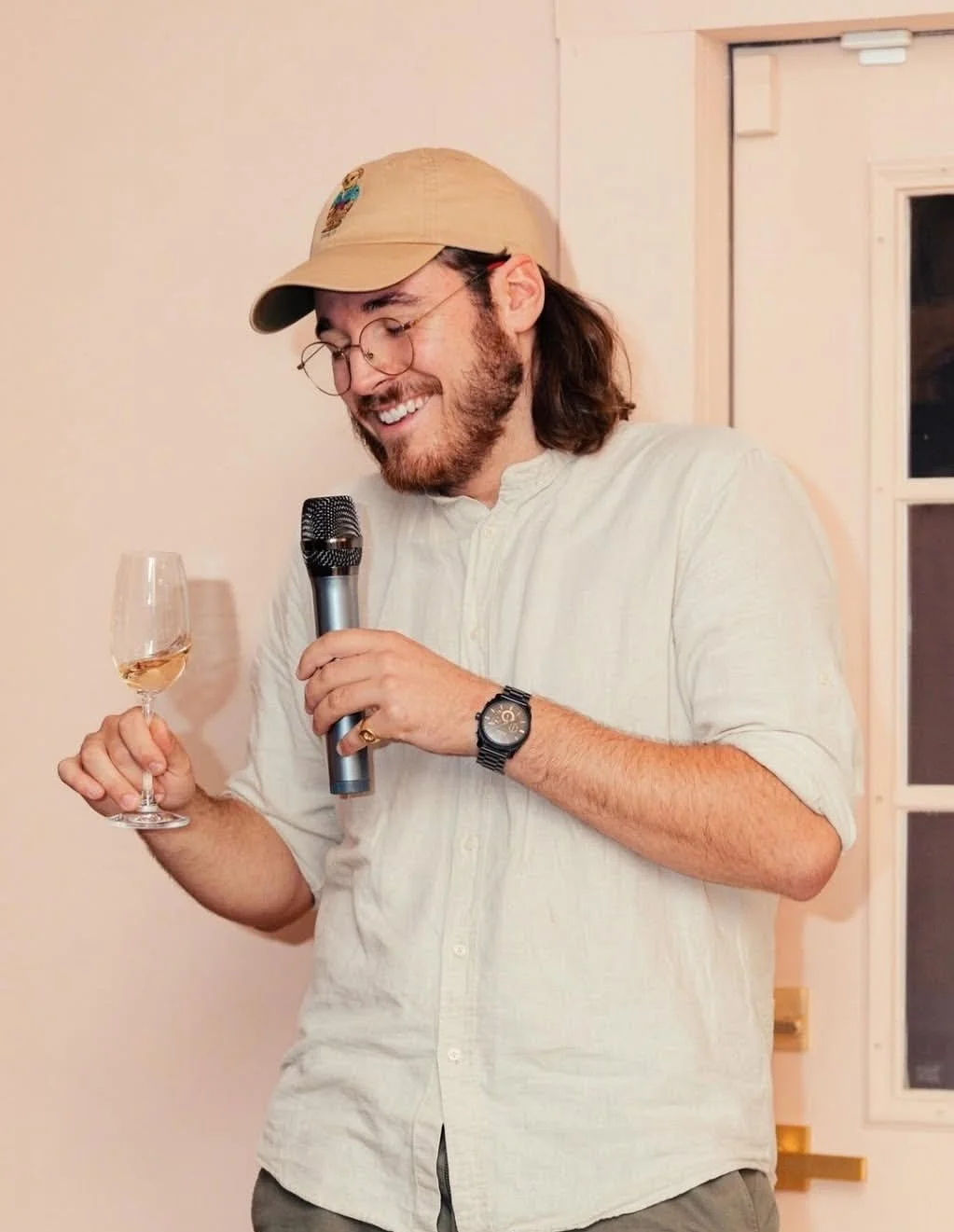 A man with glasses, a beard, and long hair, wearing a beige cap and a white shirt, is smiling and holding a microphone in one hand and a glass of white wine in the other, standing indoors near a window.