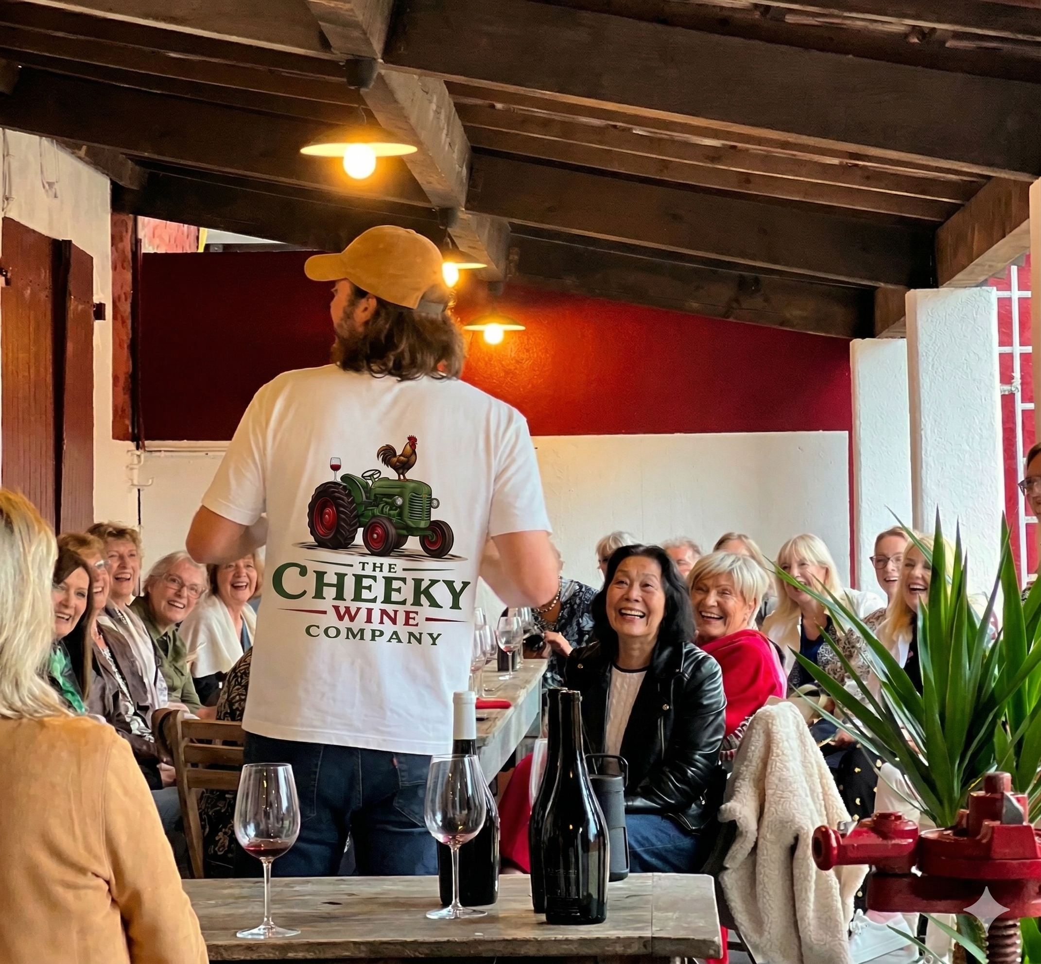 A man wearing a white t-shirt with a green tractor and a rooster on the back, and a yellow cap, serving wine to a group of people gathered around a long wooden table in a cozy restaurant with a red wall and wooden ceiling beams.